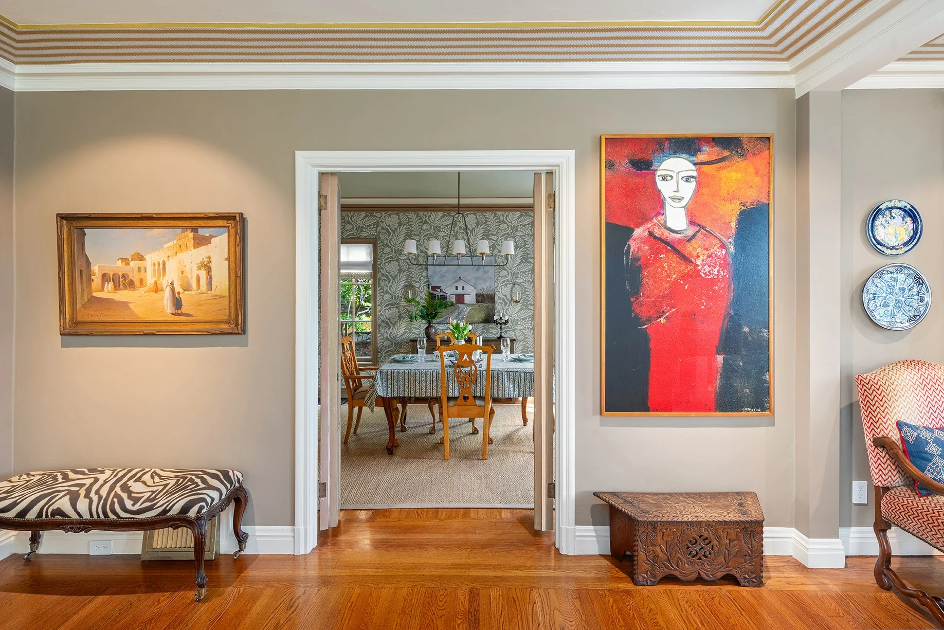 View of a living room with beige walls, large colorful paintings, and a doorway leading to a dining room with a table, chairs, and wallpapered walls.