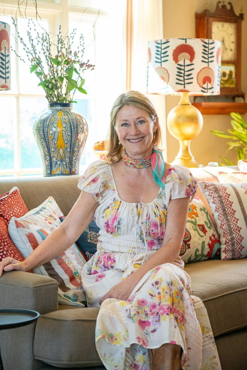 A woman with light brown hair, wearing a floral dress and a colorful scarf, sitting on a beige couch in a well-lit living room with patterned pillows, a large colorful vase with flowers, a yellow table lamp, and a wooden grandfather clock in the background.