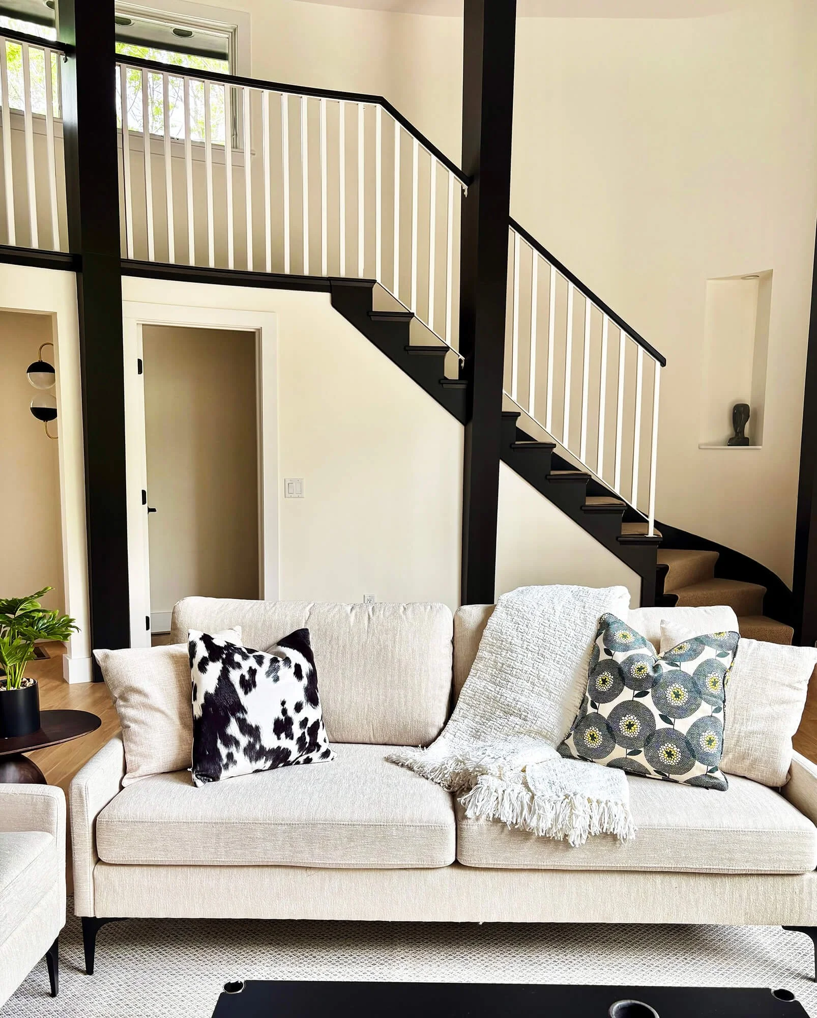Living room with a beige sofa, three decorative pillows (one with a black and white cow pattern, another with a grey and yellow floral pattern, and the third with a plain beige cover), a white throw blanket, and a small side table with a potted plant. In the background, there is a staircase with black and white railings, a wall with two rectangular windows, and a niche with a sculpture inside.