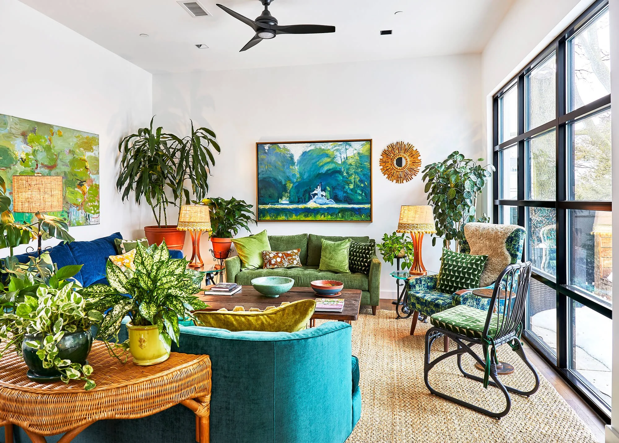 A bright, colorful living room with large windows, green and blue sofas, various patterned pillows, artwork on white walls, and numerous indoor plants. Two lamps with woven shades sit on side tables.