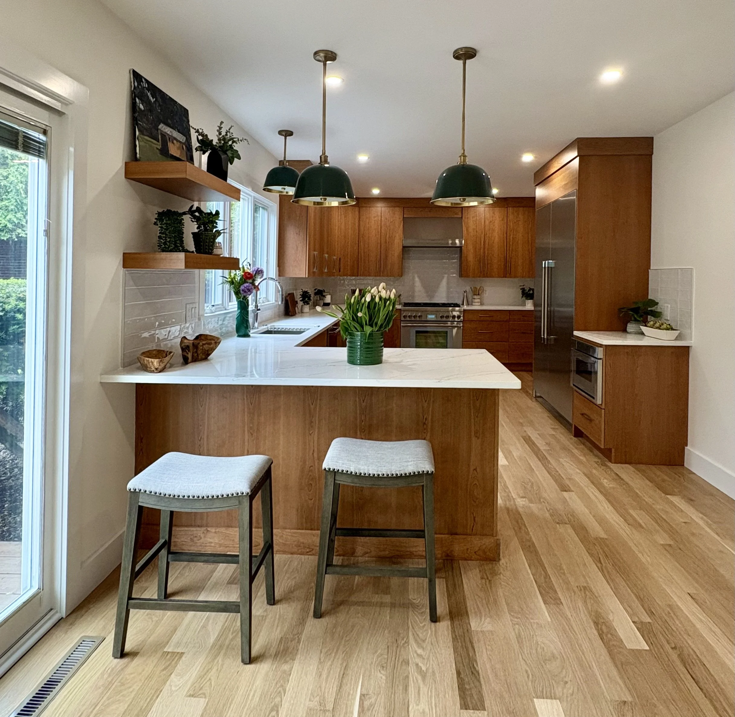A kitchen with wooden cabinets, a white marble island, pendant lights, potted plants, and a window with a sliding door.
