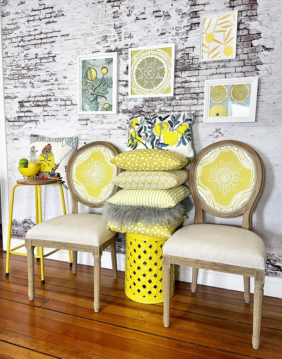 Decorative indoor wall with framed botanical artwork, two wooden chairs with yellow and white floral upholstery, a stack of yellow-patterned pillows, a yellow side table with a bowl, mug, and small figurine, and a white brick wall background.