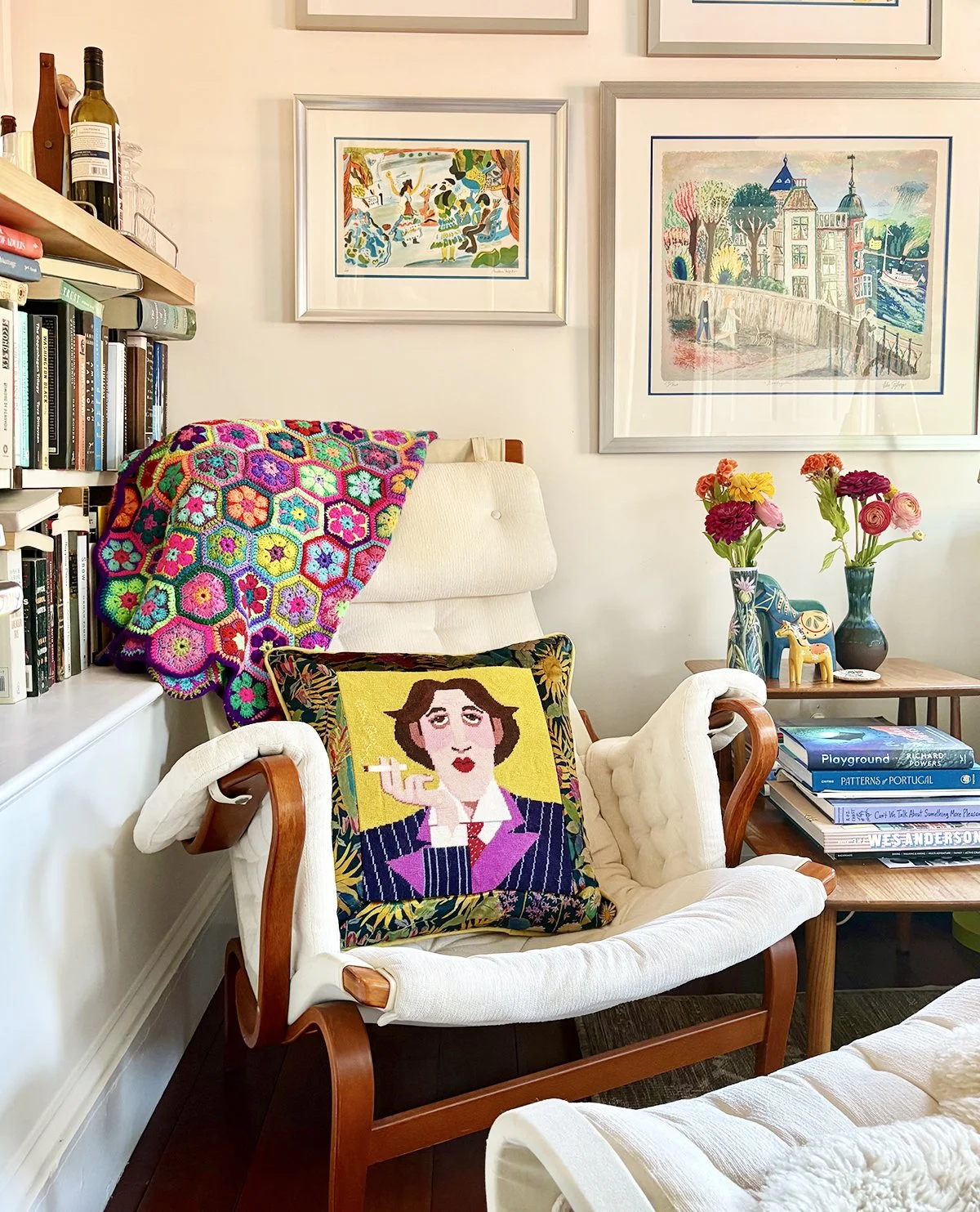 A cozy living room corner with an armchair that has a colorful needlepoint pillow depicting a woman holding a cigarette. A crocheted blanket with multicolored floral hexagons is draped over the back of the chair. Next to the chair is a small wooden side table holding books, vases with flowers, and decorative objects. The wall features framed artwork including vibrant landscape and figurative paintings. A bookshelf filled with books is partially visible on the left.