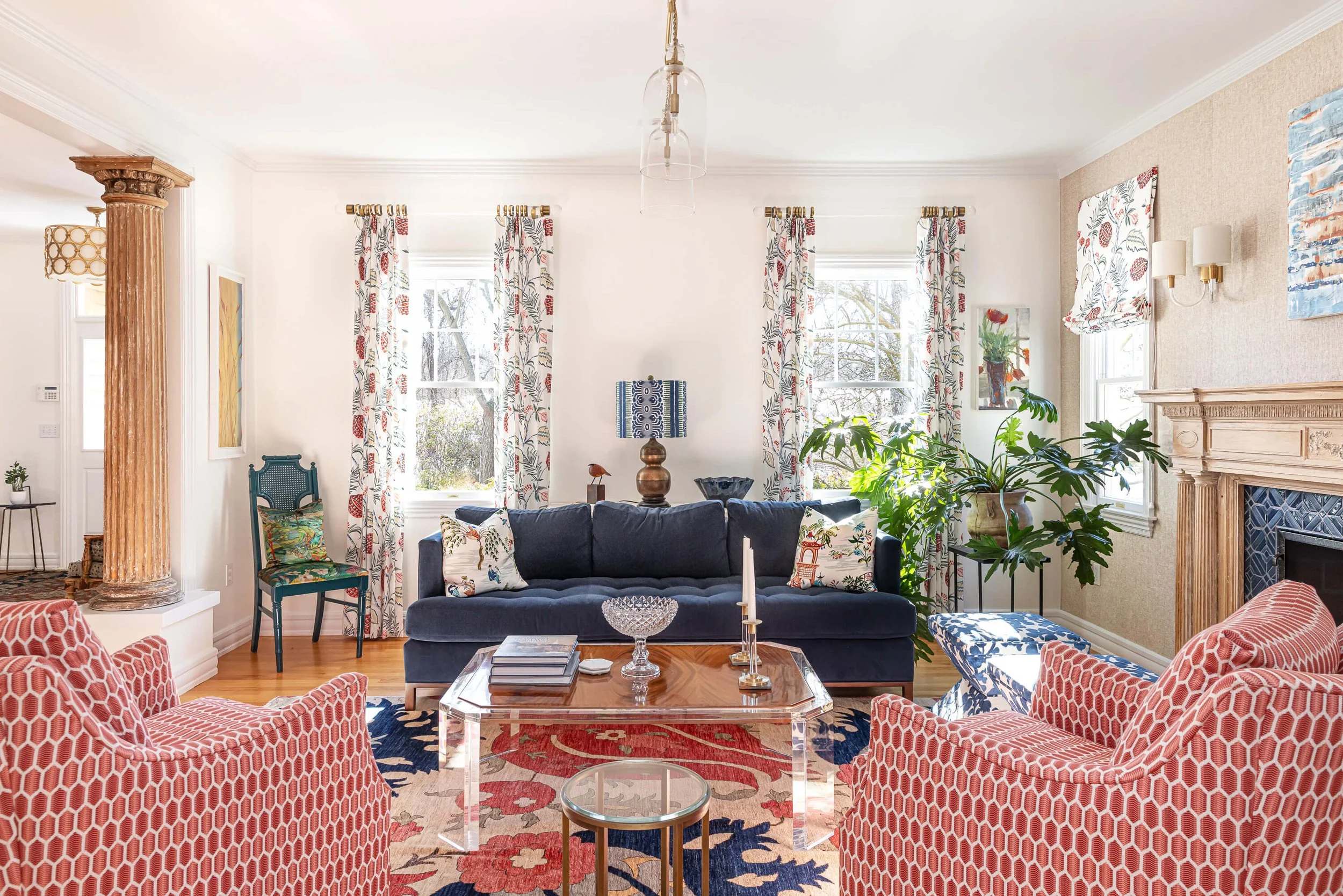 A living room with a navy sofa with patterned pillows, a glass coffee table, red patterned armchairs, large windows with floral curtains, a fireplace with a decorative mantel, and various decorative items and plants.