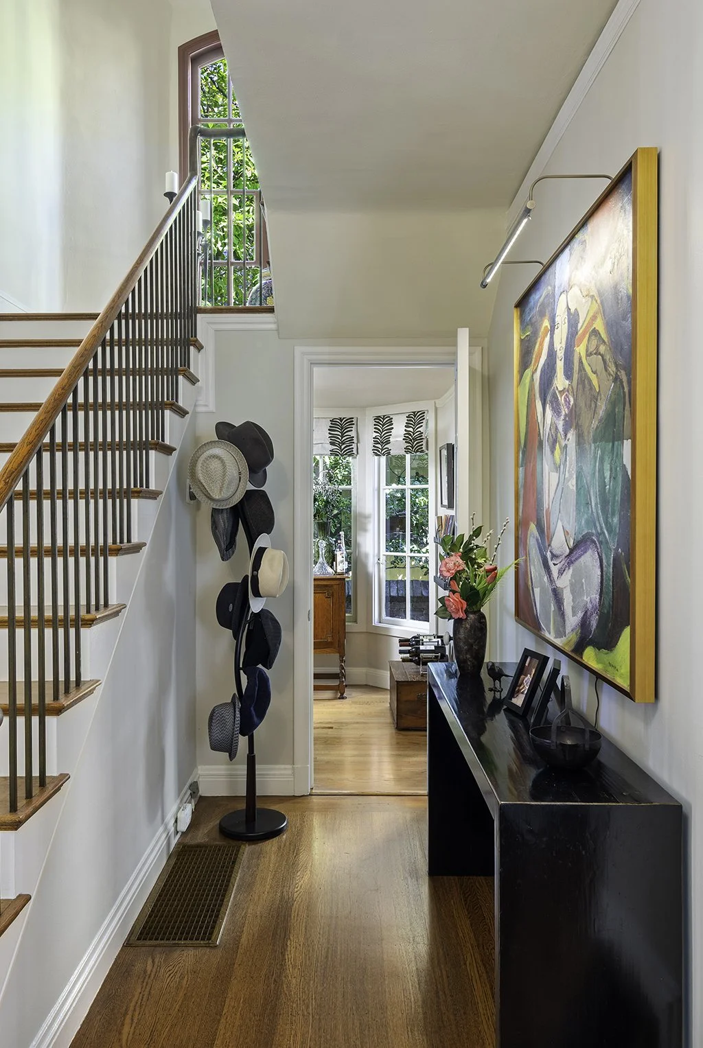 Entryway of a home with a staircase on the left, hats hanging on a stand, a black console table with a vase of pink flowers and framed photos, an abstract painting on the wall, and a view into a sunlit room with bay windows and greenery outside.