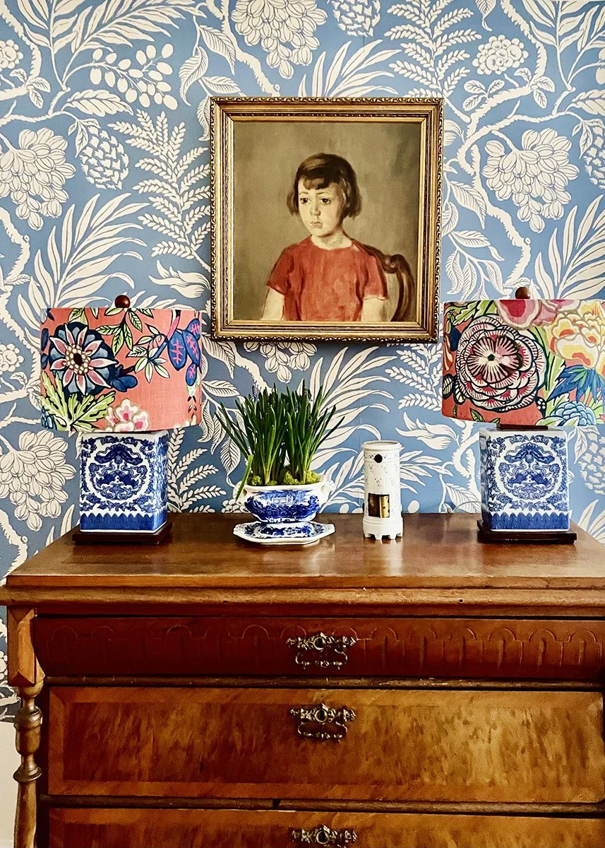A wooden dresser with two colorful floral lamps, a potted green plant, and a white humidifier on top, against a blue and white floral wallpaper background. A framed portrait of a young girl in a red shirt hangs above.