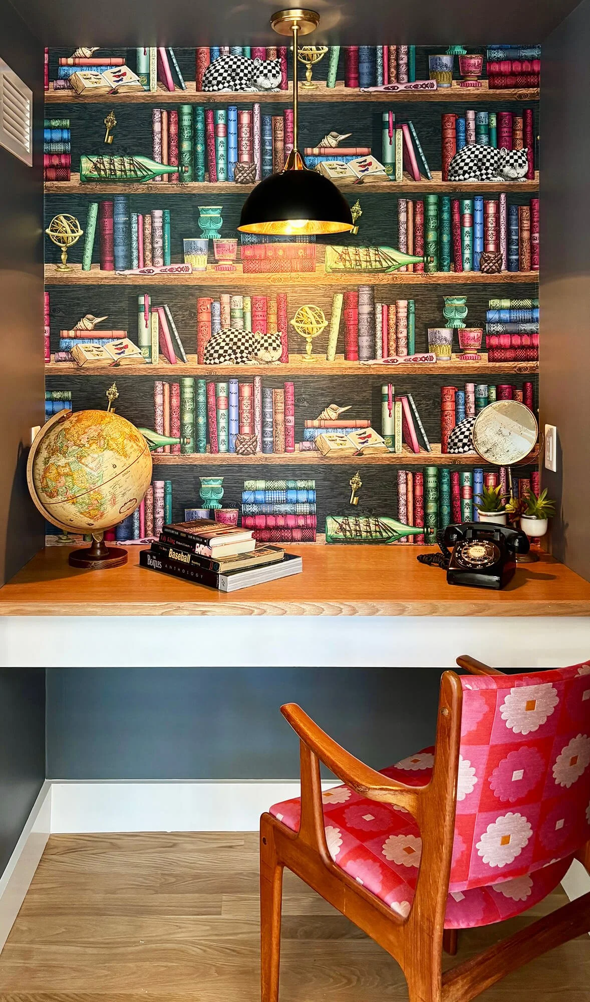 A cozy reading nook with a vibrant bookshelf wallpaper, a wooden desk with a globe, books, an old-fashioned rotary phone, and small plants. A pink patterned armchair with wooden arms is in the foreground.