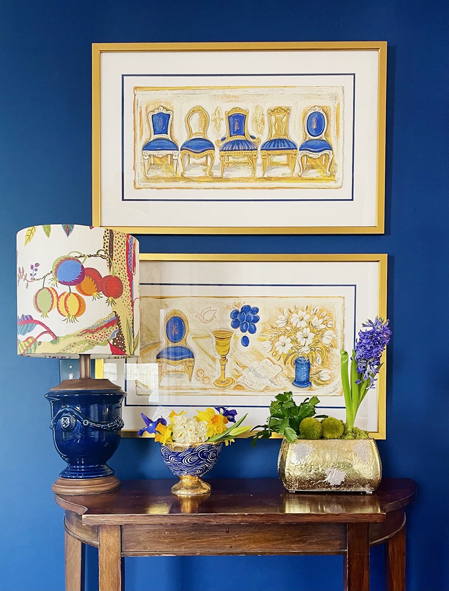 Decorative table with colorful flowers in a blue and gold bowl, a dark blue ceramic urn, and a gold-colored vase with purple hyacinths and green foliage against a blue wall. Two framed artworks of antique chairs and still life objects hang above the table.