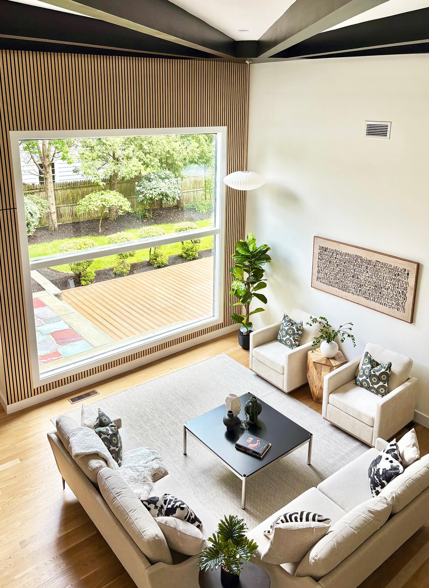 A modern living room with a large beige sofa, a black coffee table with books and decorative items, and an armchair with patterned pillows. There are plants, a white wall with artwork, and large windows showing a lush backyard with trees and a wooden deck.
