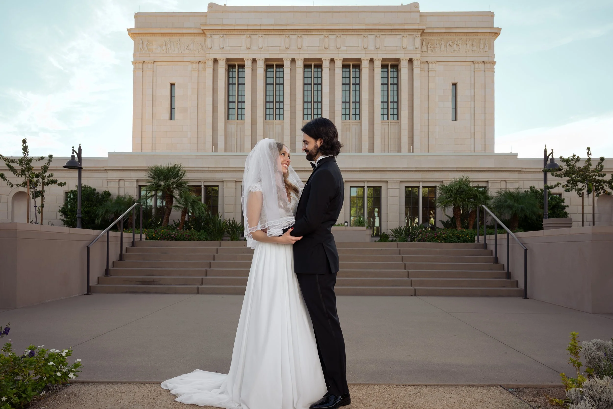 A bride and groom standing face to face in front of the mesa mormon church temple with stairs, palm trees, and lampposts, holding hands.