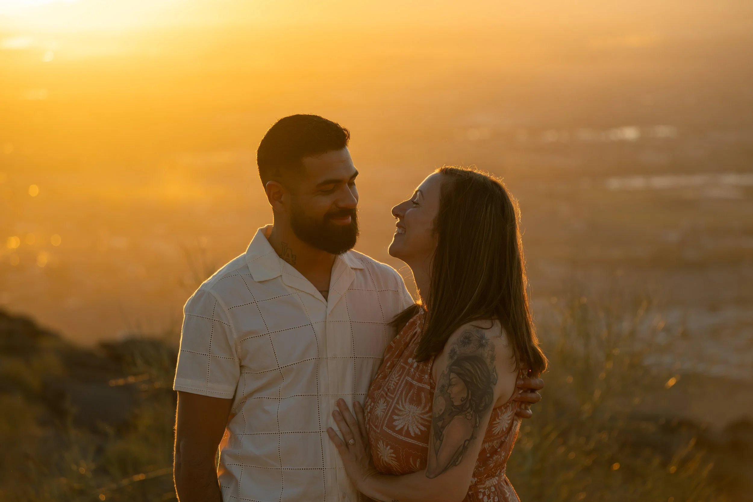 A couple standing close together on a mountain during sunset, smiling and looking into each other's eyes.