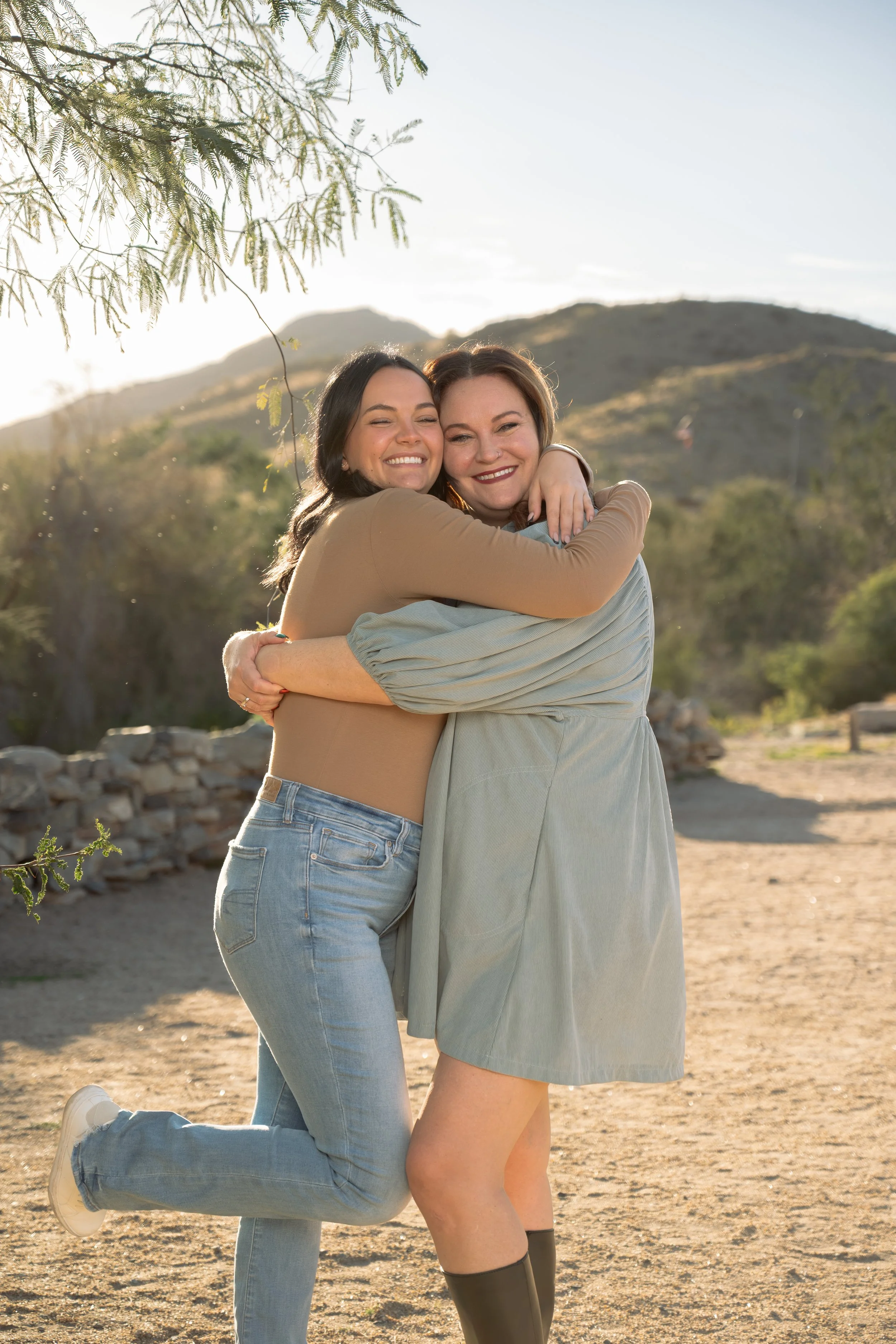 Two women hugging and smiling outdoors near mountains and trees in warm sunlight.
