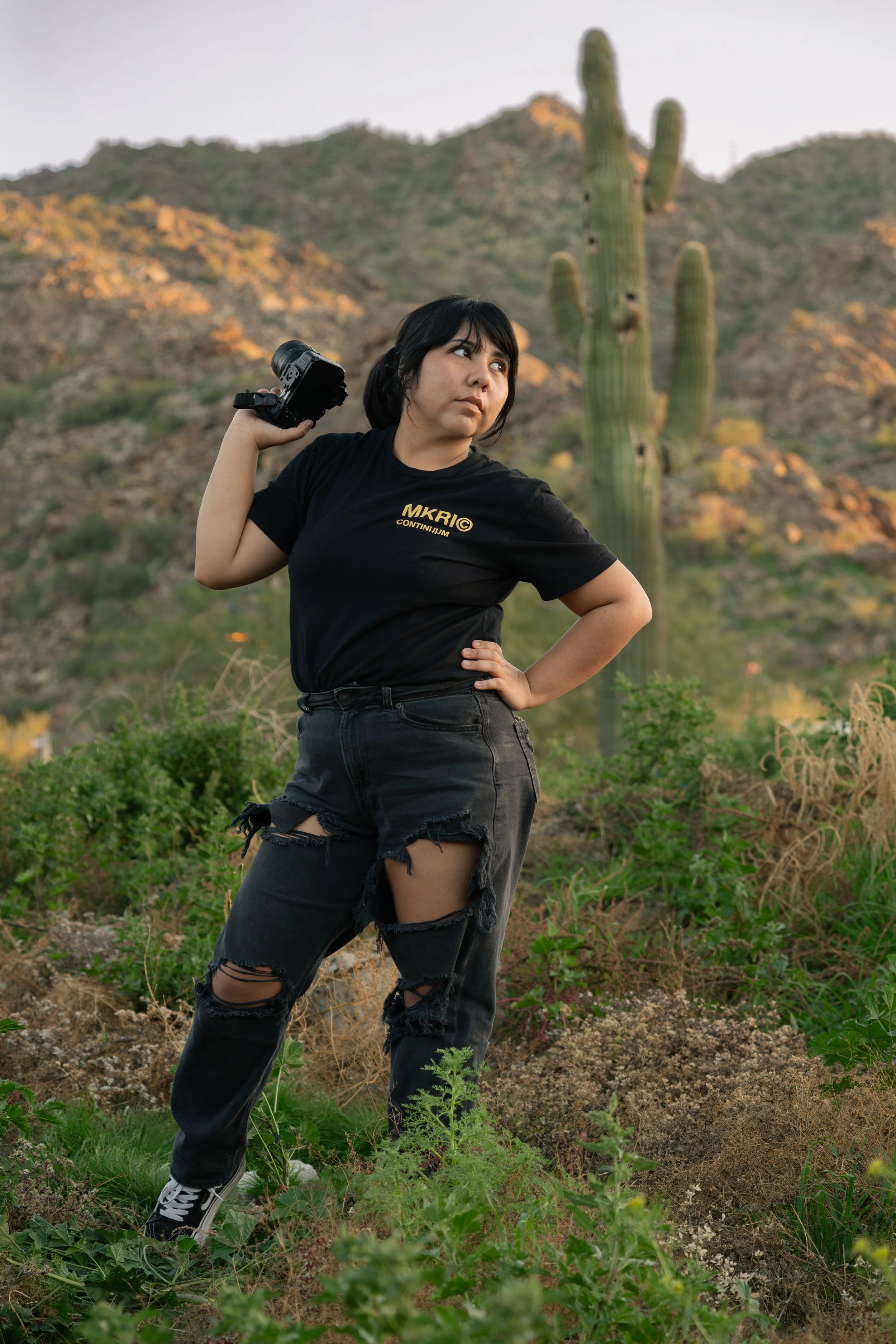 Young woman standing outdoors in a scrubby, desert landscape with hills and a tall saguaro cactus in the background. She is holding a camera behind her head with her right hand, standing with her left hand on her hip, wearing a black t-shirt with yellow text, ripped black jeans, and sneakers.