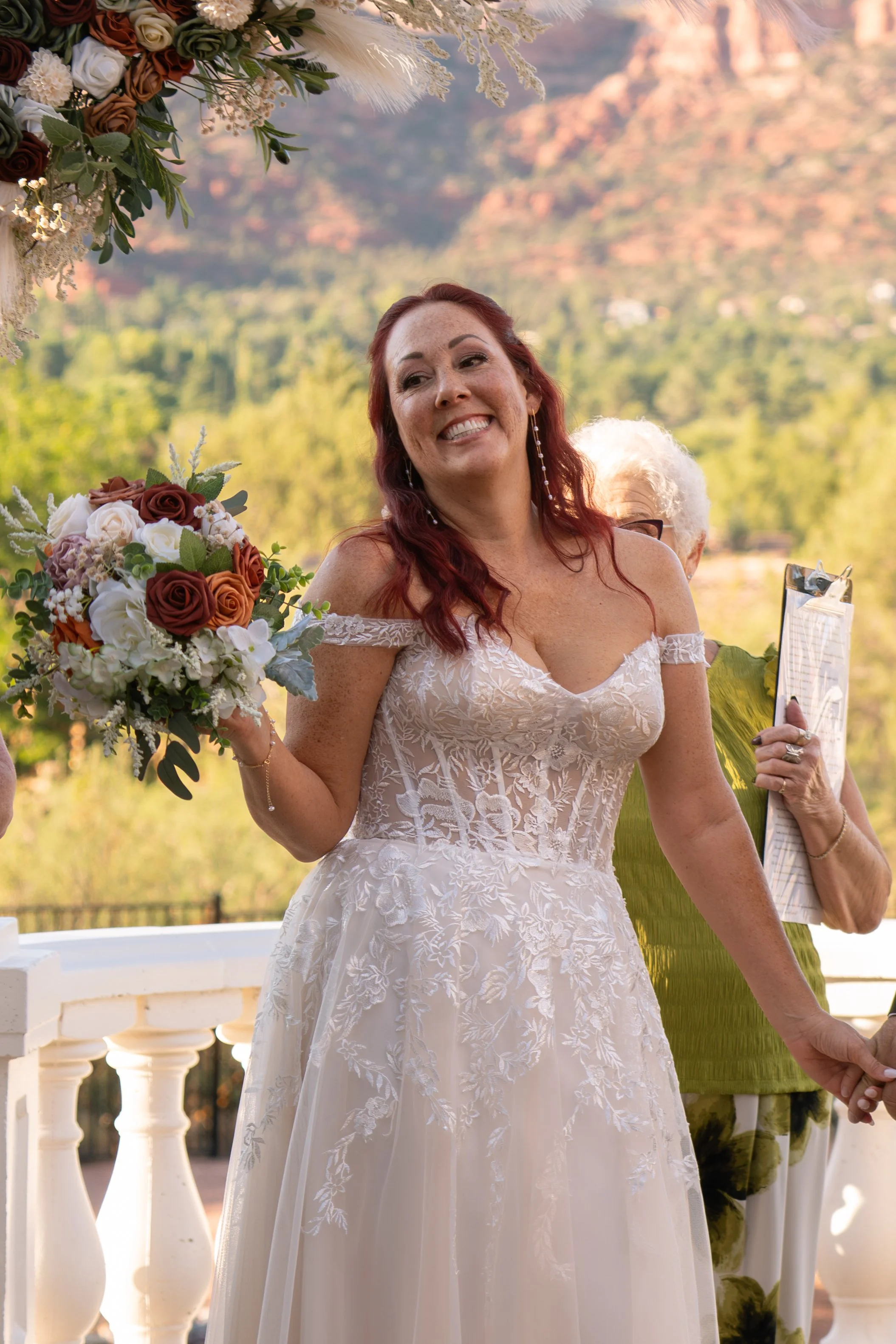 A bride in a white lace wedding dress holding a bouquet of flowers, smiling outdoors with mountains and trees in the background.