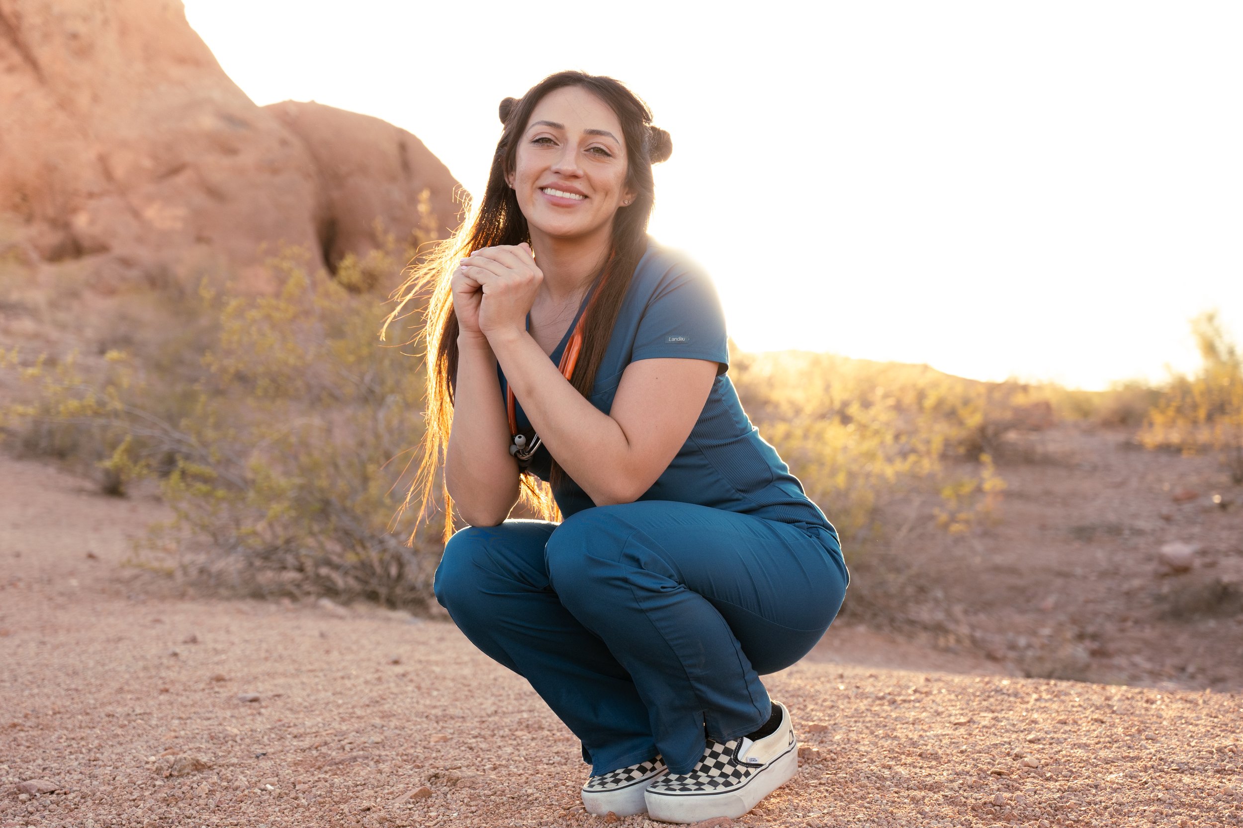 A woman in medical scrubs crouches on a rocky desert terrain, smiling at the camera with her hands clasped near her chin, under a bright sunset sky.