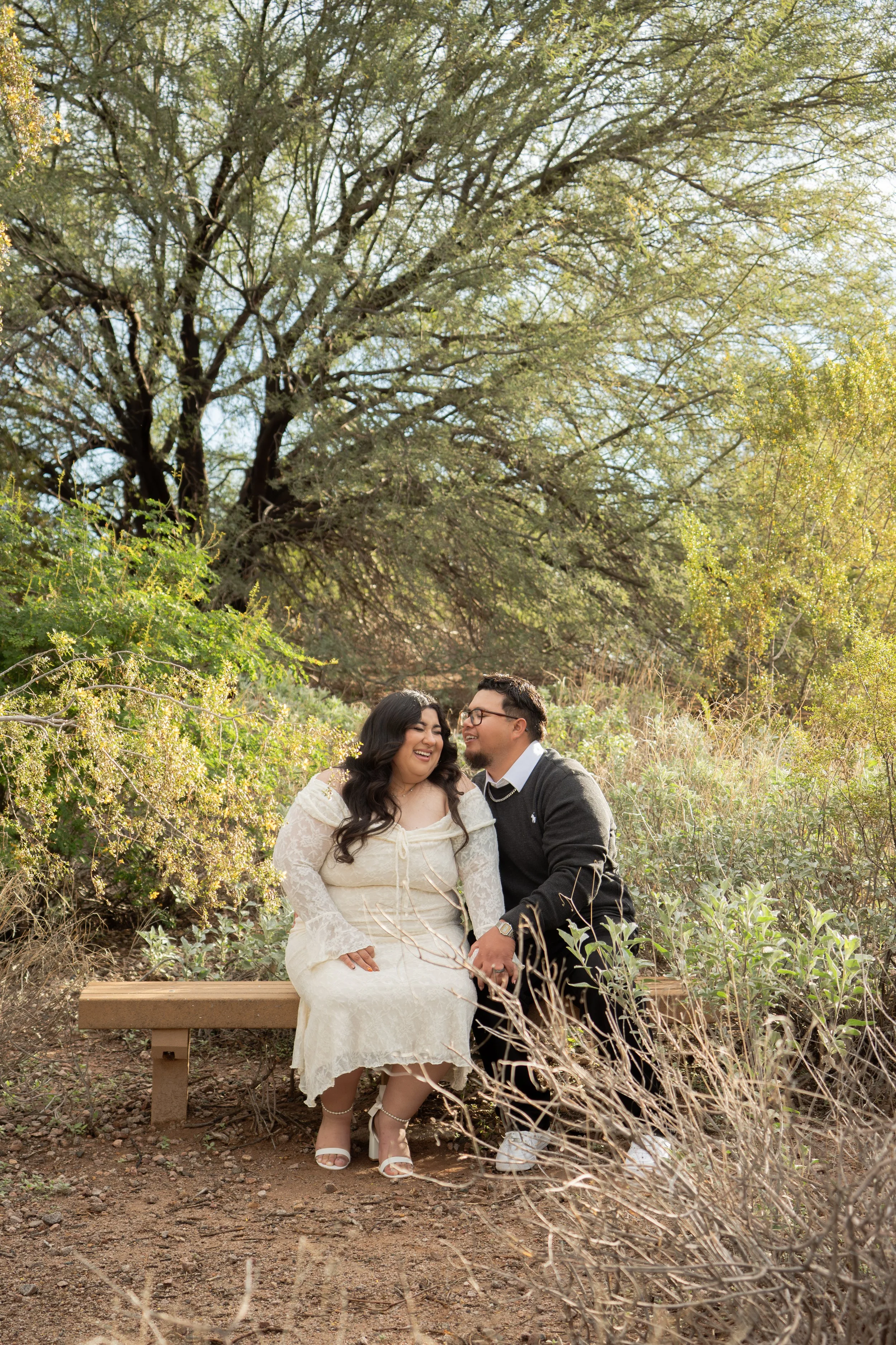 A joyful couple sits on a bench outdoors surrounded by greenery and trees, sharing a happy moment.