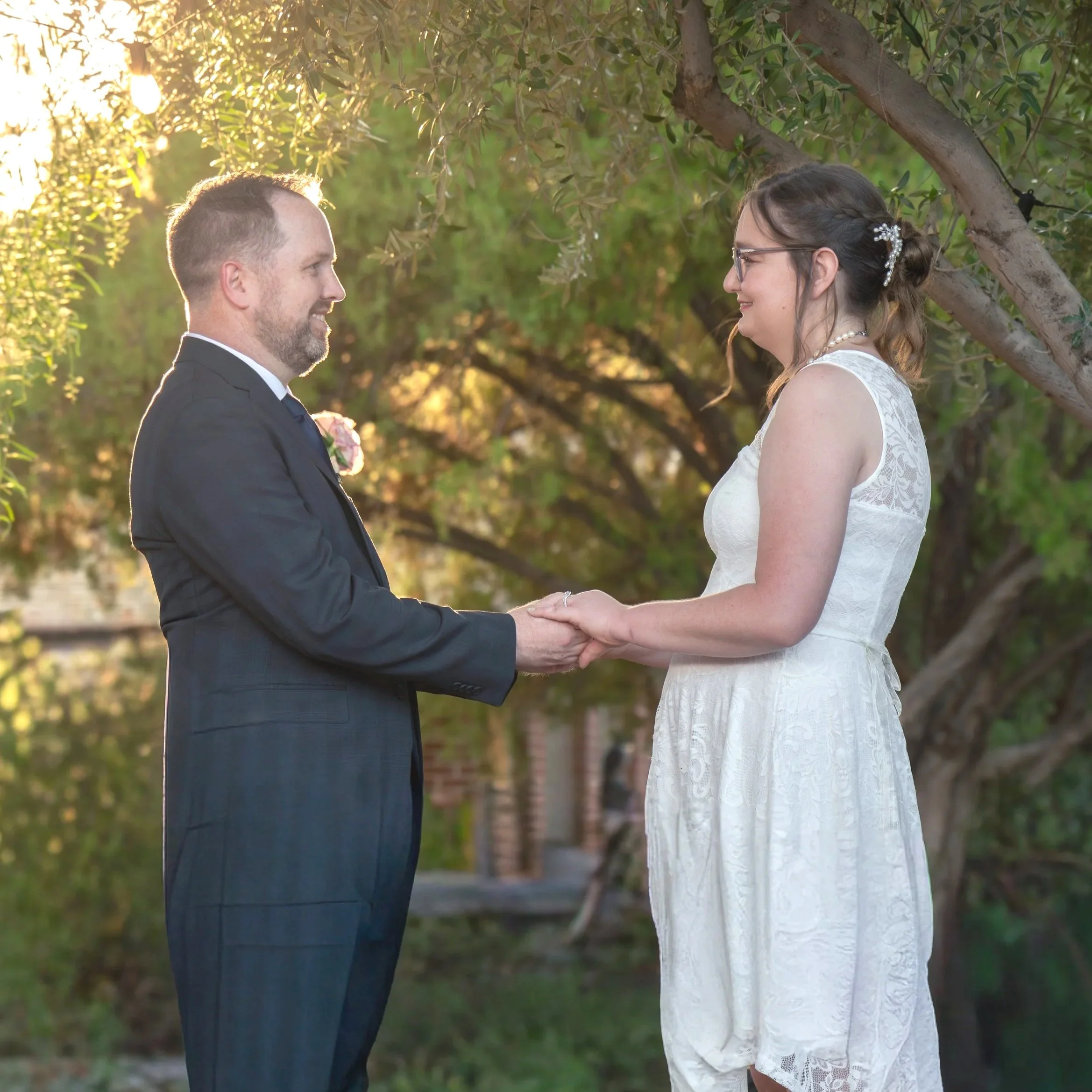A couple holding hands and smiling at each other outdoors during a wedding, with trees and warm sunlight in the background.