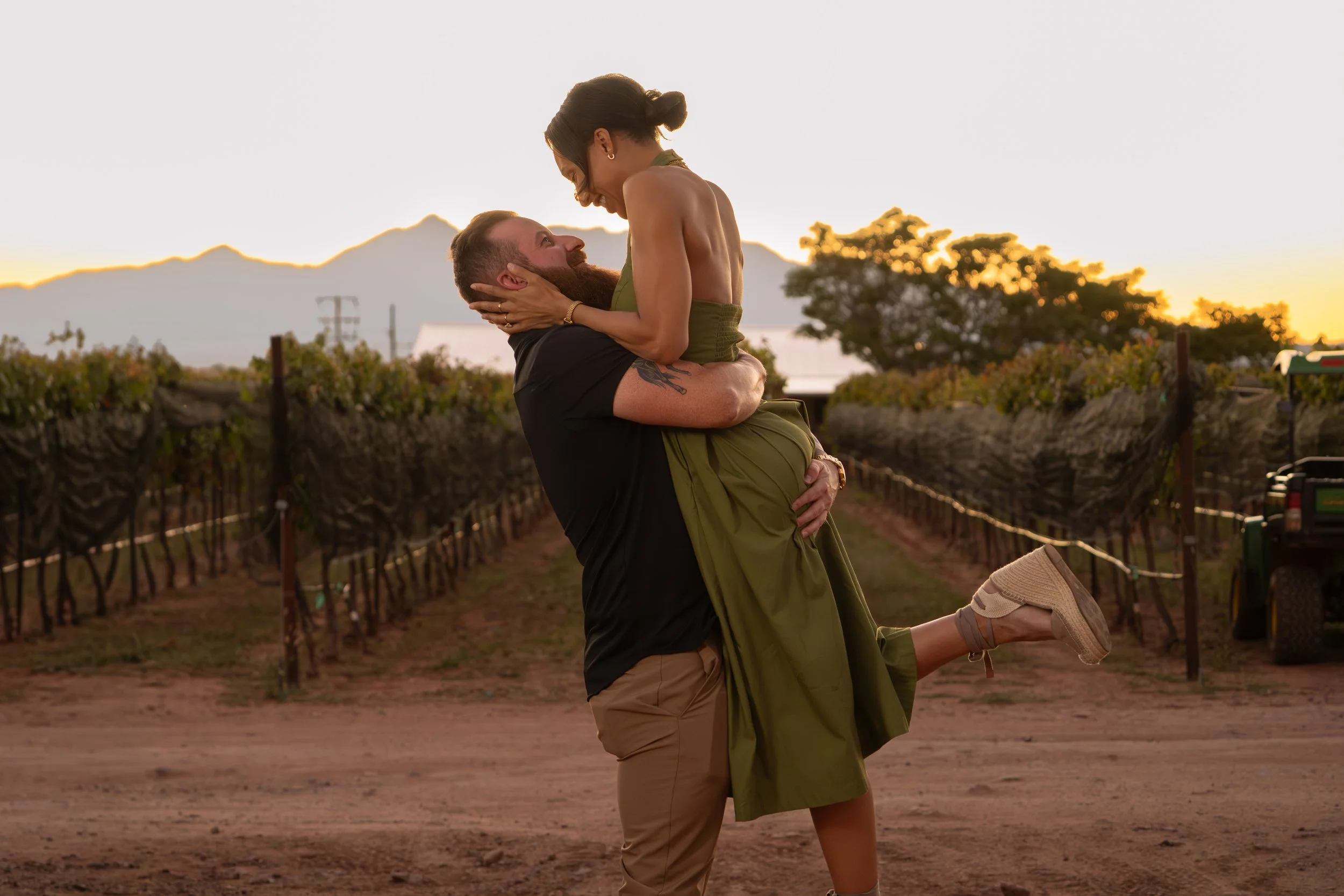 A man and woman kissing outdoors at sunset with a blurred background of green foliage.