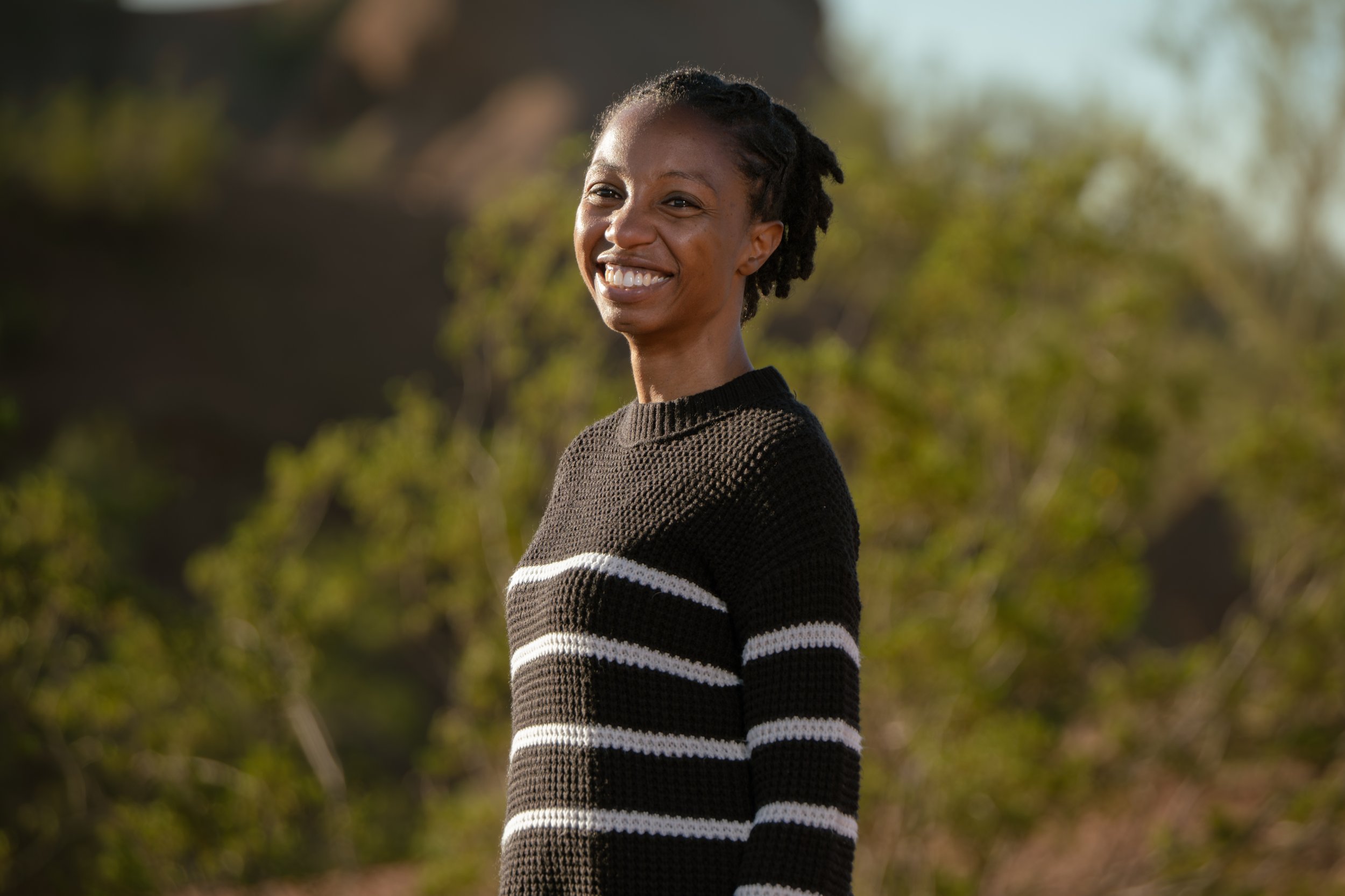 A man and a young boy, both wearing black Phoenix Suns t-shirts, are outdoors in a park. The boy is sitting on a small orange and black bike with a purple helmet, and the man is kneeling next to him, smiling. The background features trees and a dirt path.