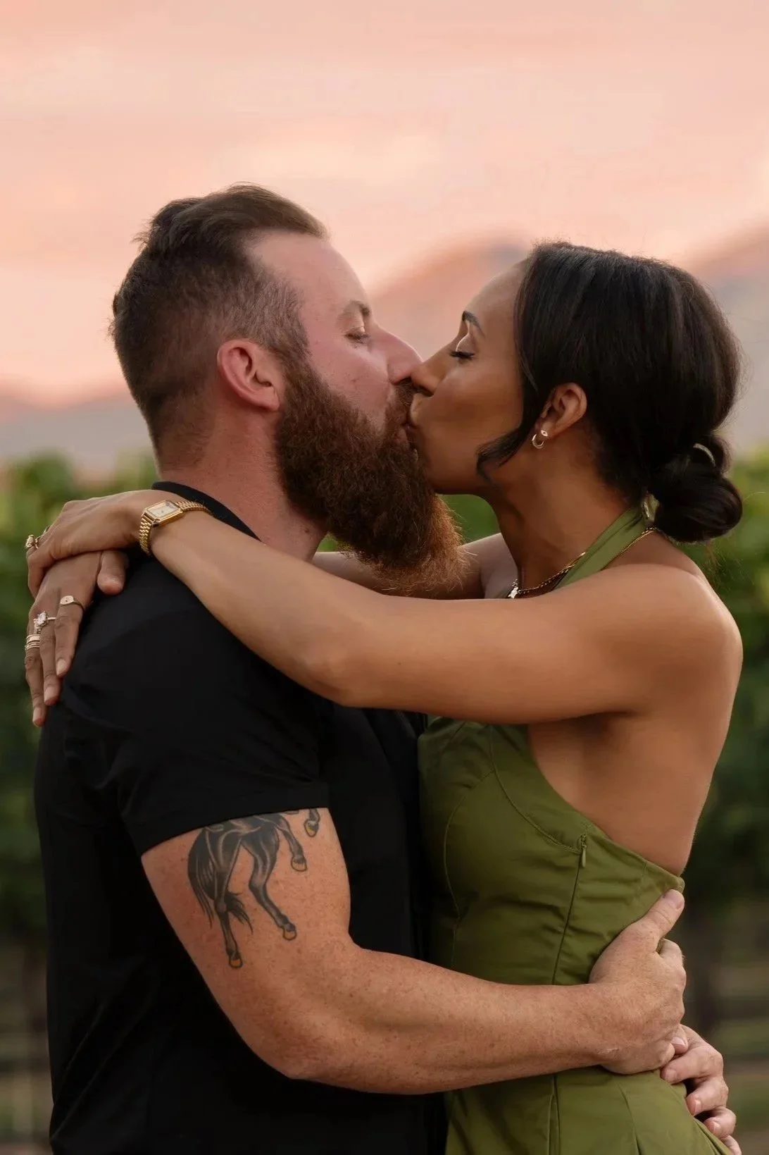 A man and woman kissing outdoors at sunset with a blurred background of green foliage.