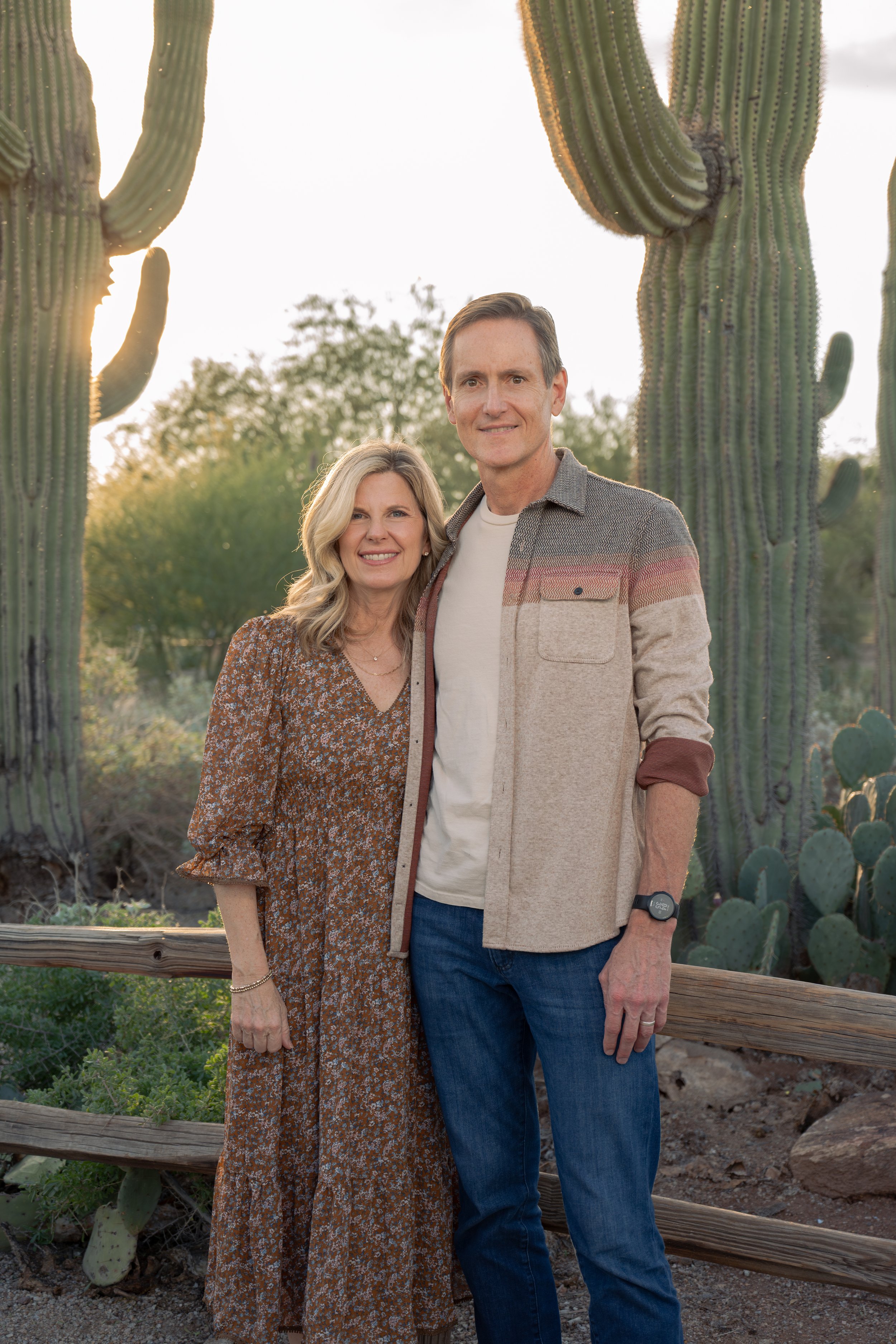 A smiling middle-aged woman and a tall middle-aged man standing close together outdoors during sunset, surrounded by large cacti and desert plants.