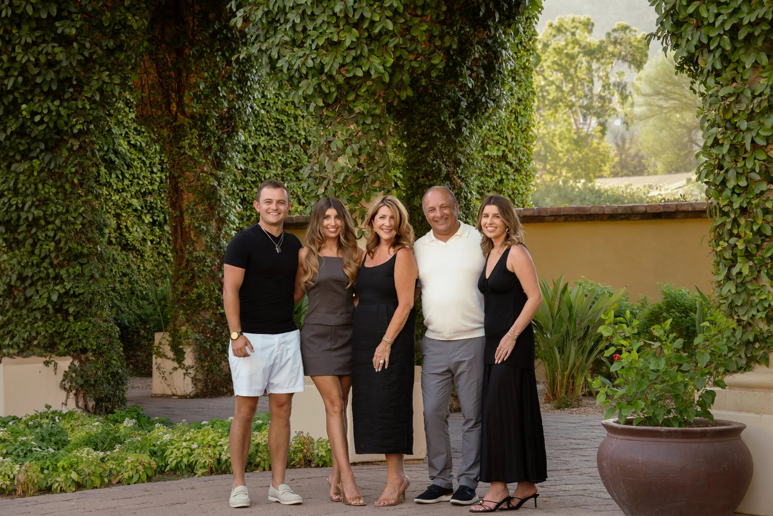 A group of five people, two men and three women, standing outdoors in front of a lush green background, smiling for the camera on a sunny day in arizona.