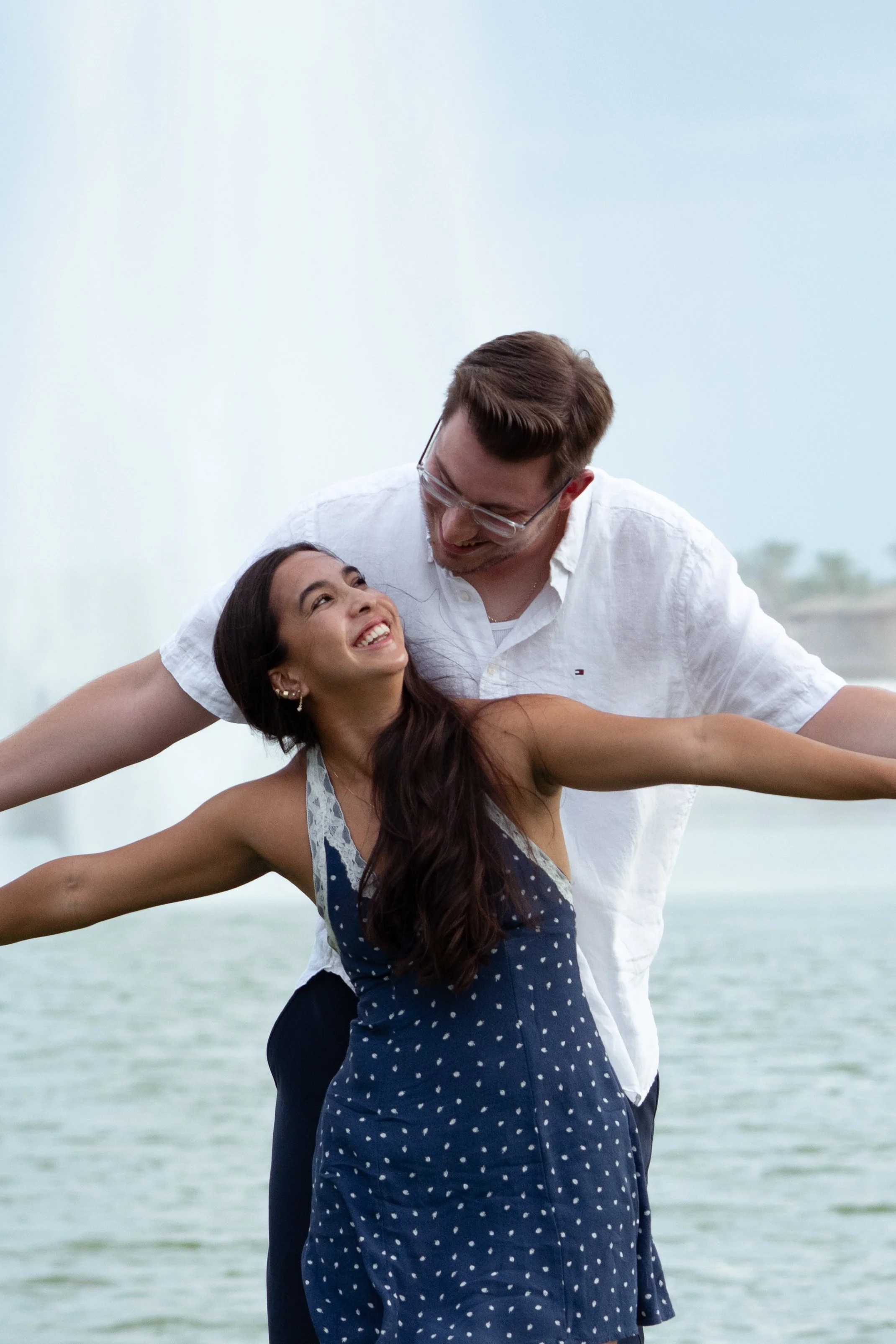 A happy couple near a waterfall, with the woman in a blue polka-dot dress and the man in a white shirt, smiling and embracing each other.