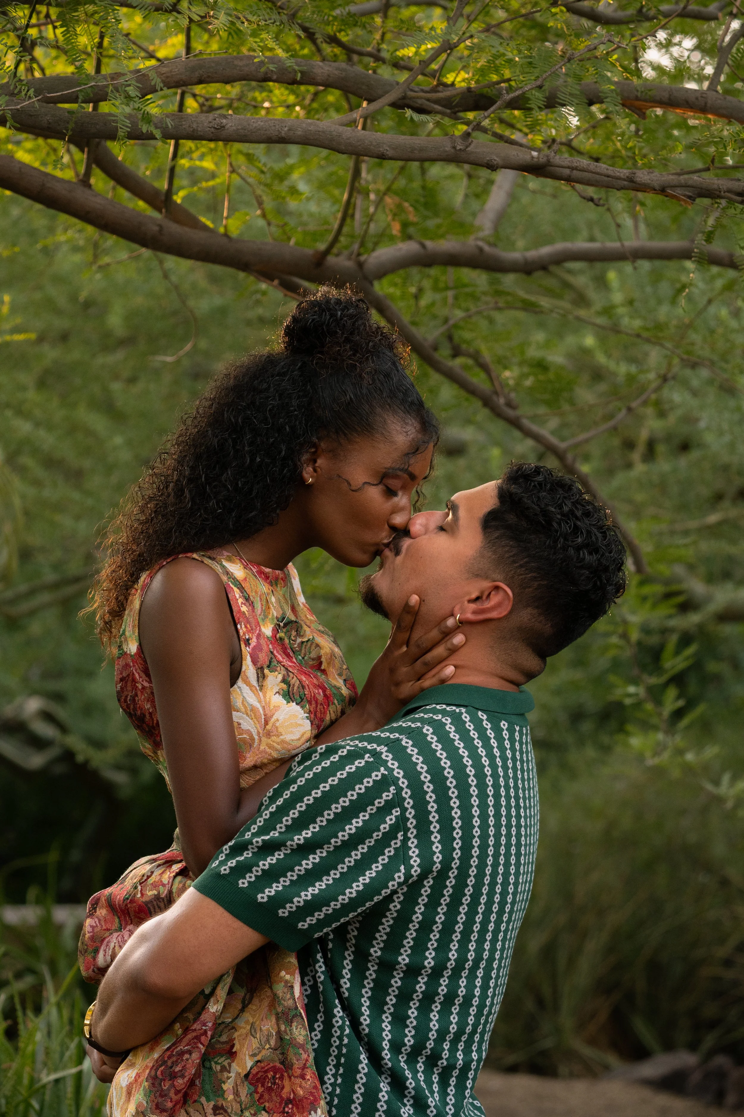 A couple sharing a kiss outdoors among trees and greenery.