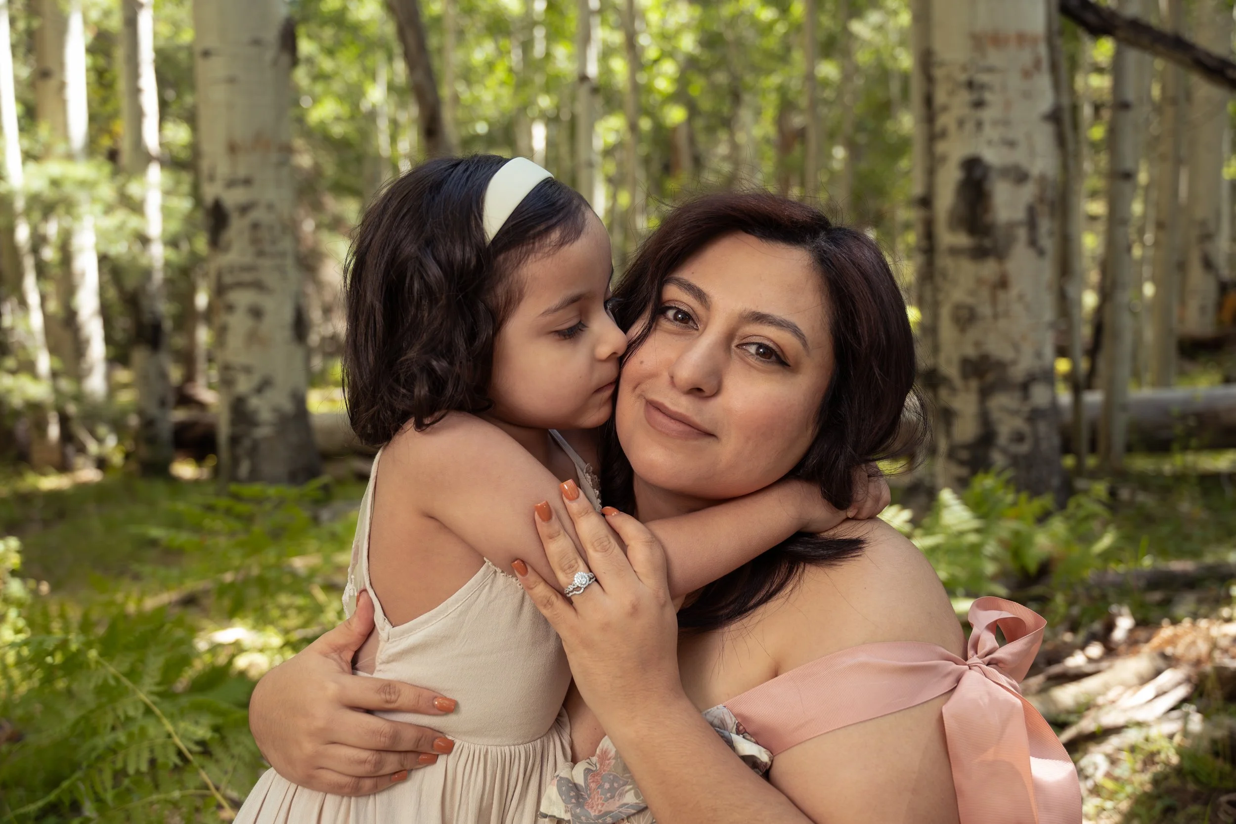 A woman and young girl hugging in a forest, with the girl kissing the woman's cheek.