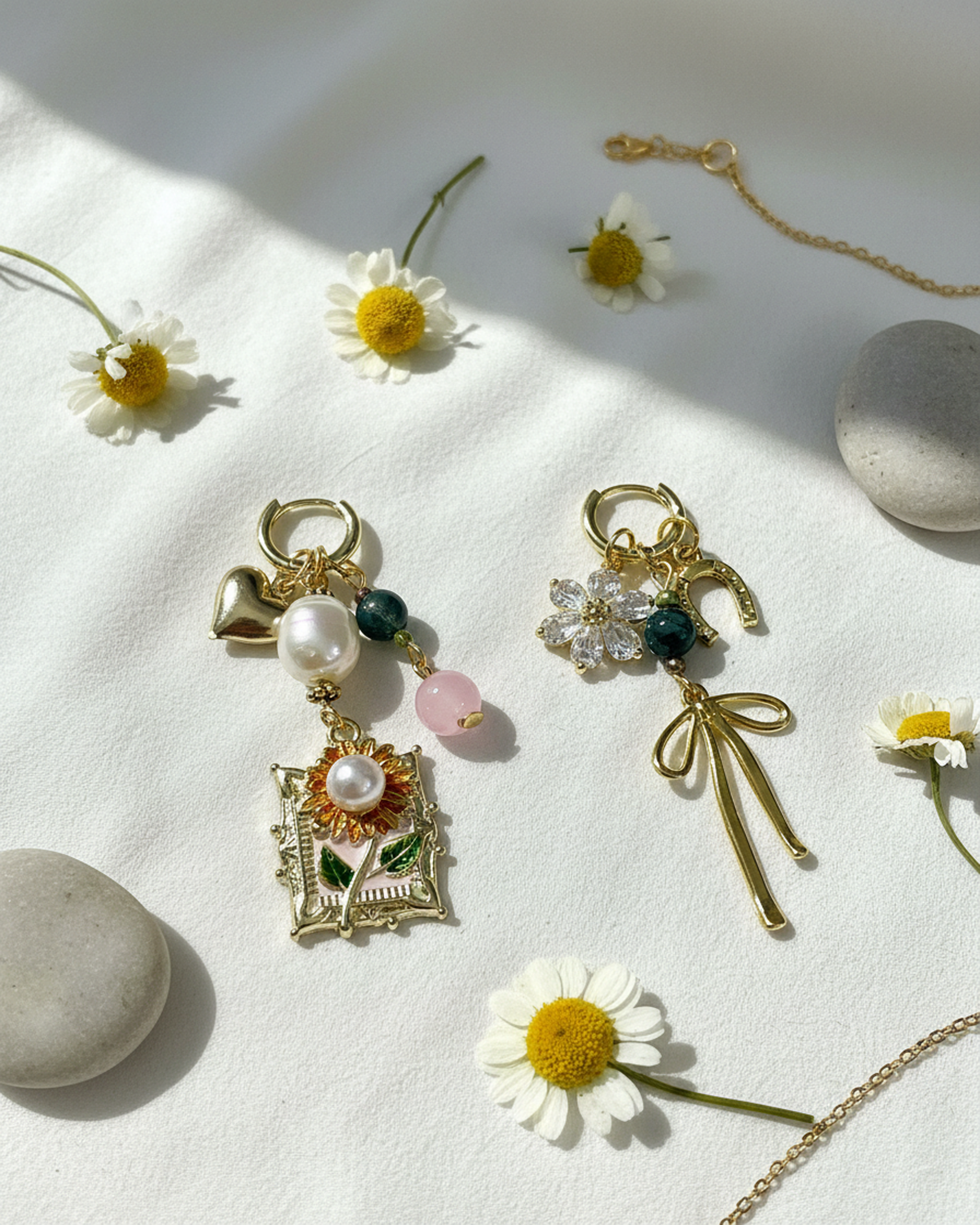 Two gold charm earrings with floral and bow designs, surrounded by small white daisies, stones, and a delicate chain on white textured paper