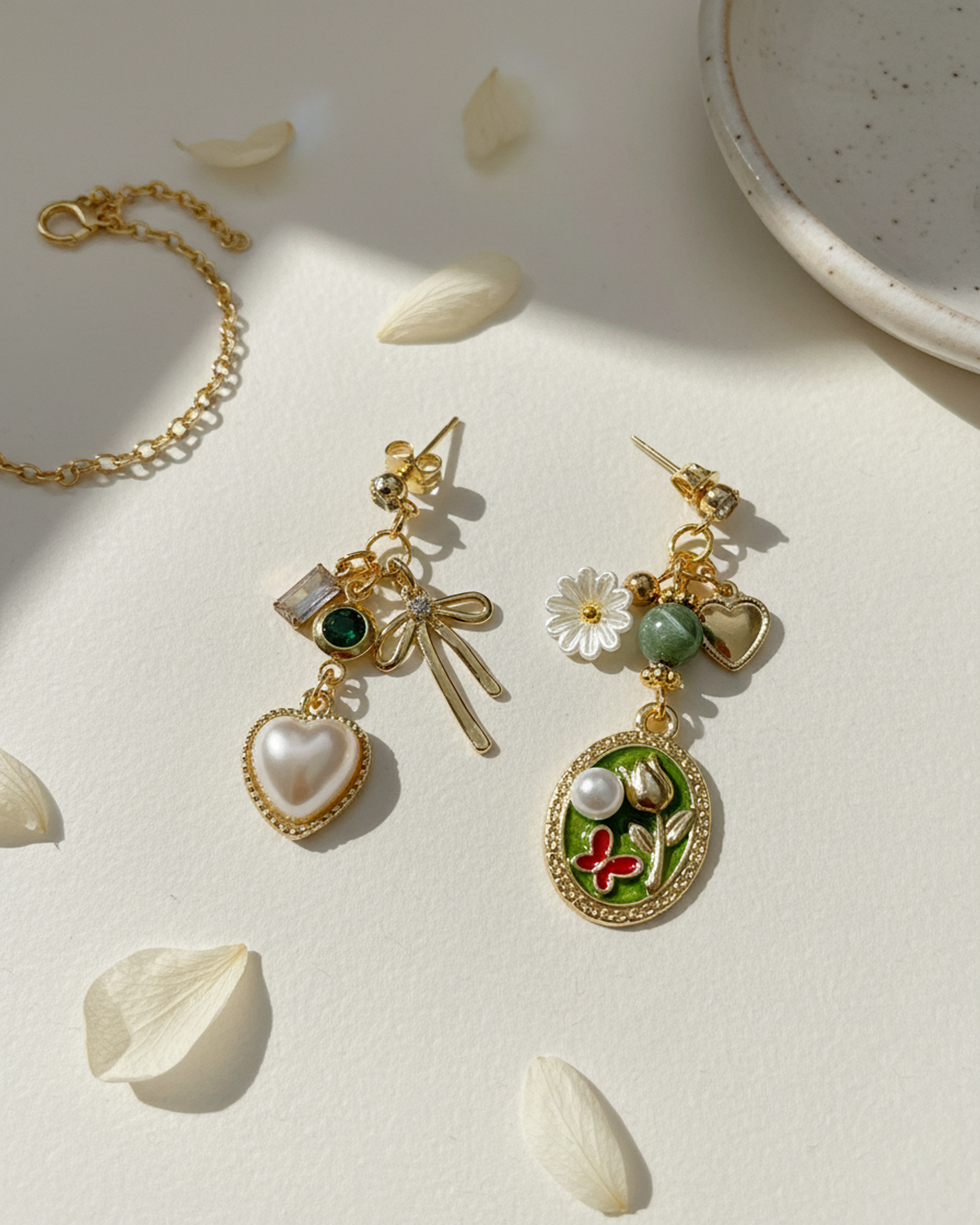 A pair of gold earrings with various charms, including a heart, flower, butterfly, and bow, on a white surface with scattered flower petals and a ceramic dish.
