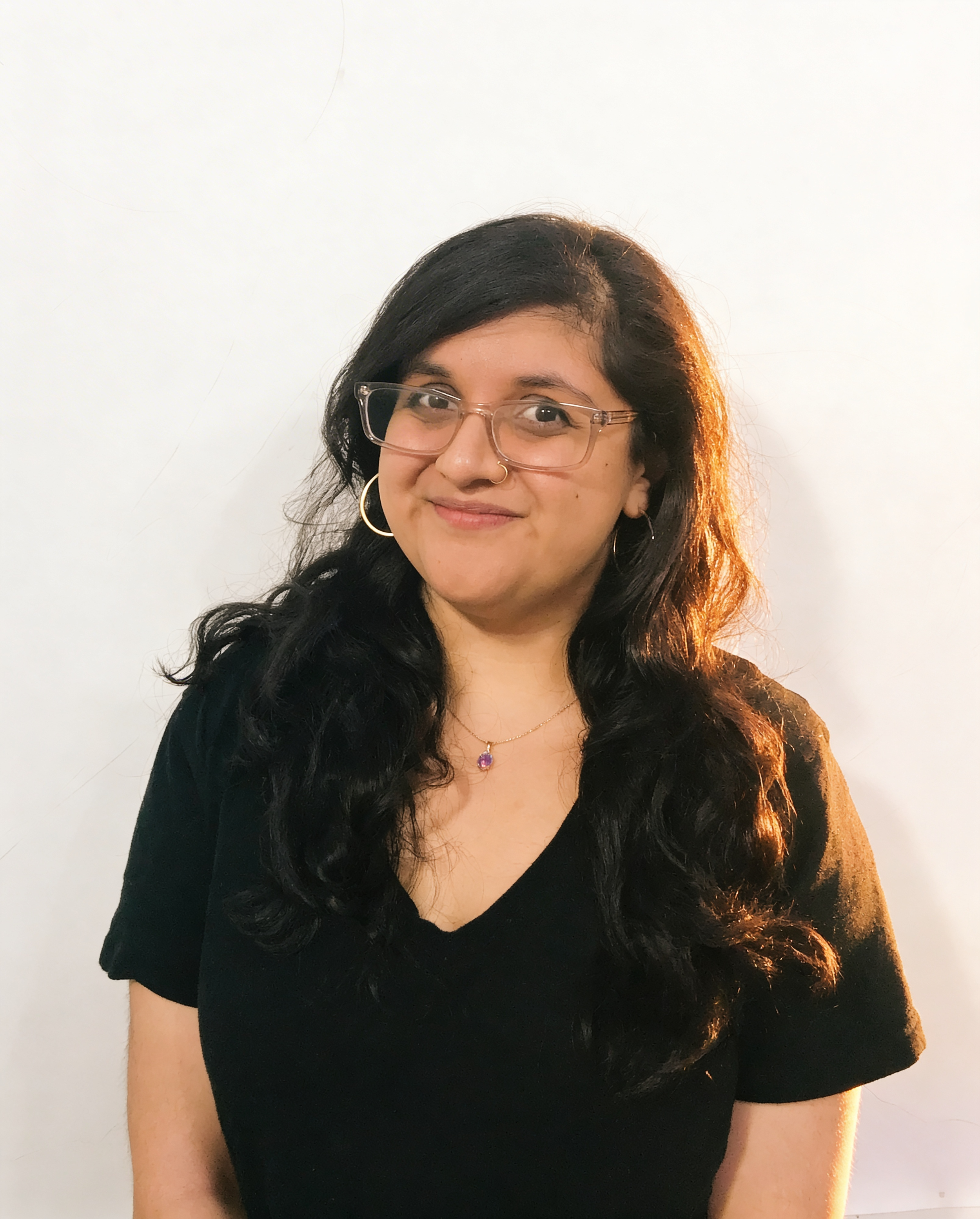 A woman with long, wavy black hair, wearing glasses, a black top, hoop earrings, a necklace with a purple pendant, and a nose ring, smiling with a plain white background.