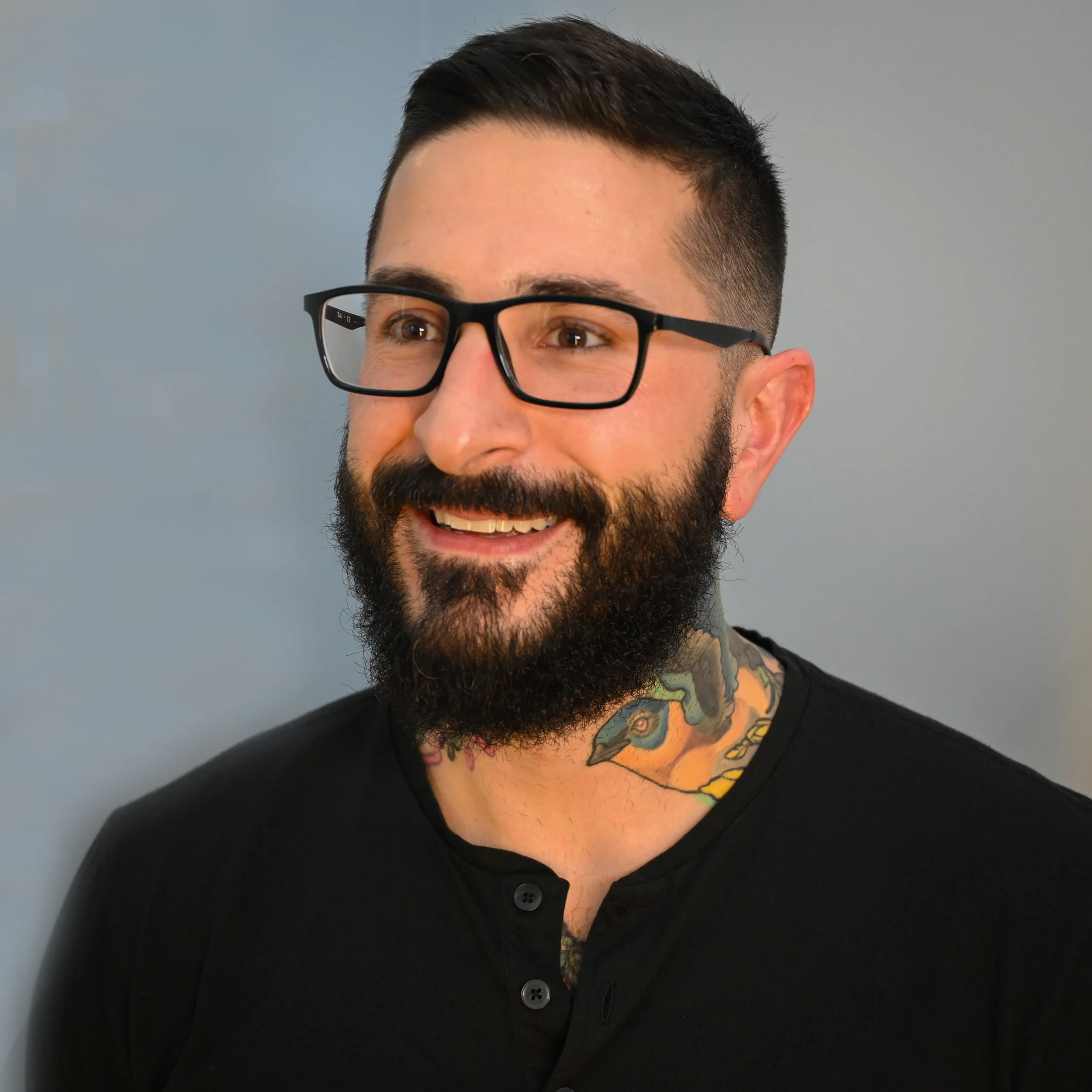 Close-up of a smiling man with a beard, glasses, and a neck tattoo, wearing a black shirt, against a gray background.