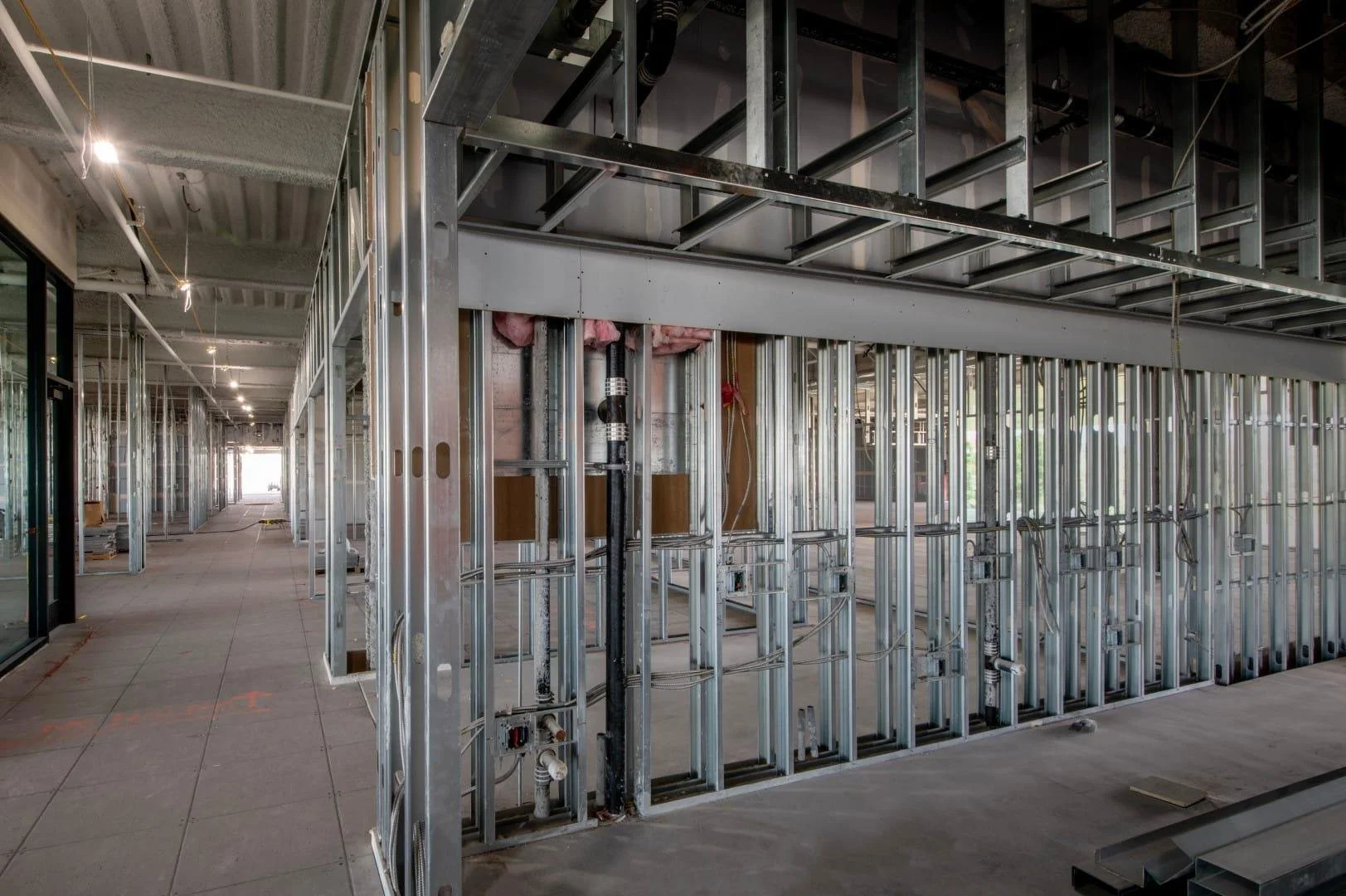 Interior of a building under construction showing metal framing for walls and ceiling, with electrical wiring and plumbing pipes installed.