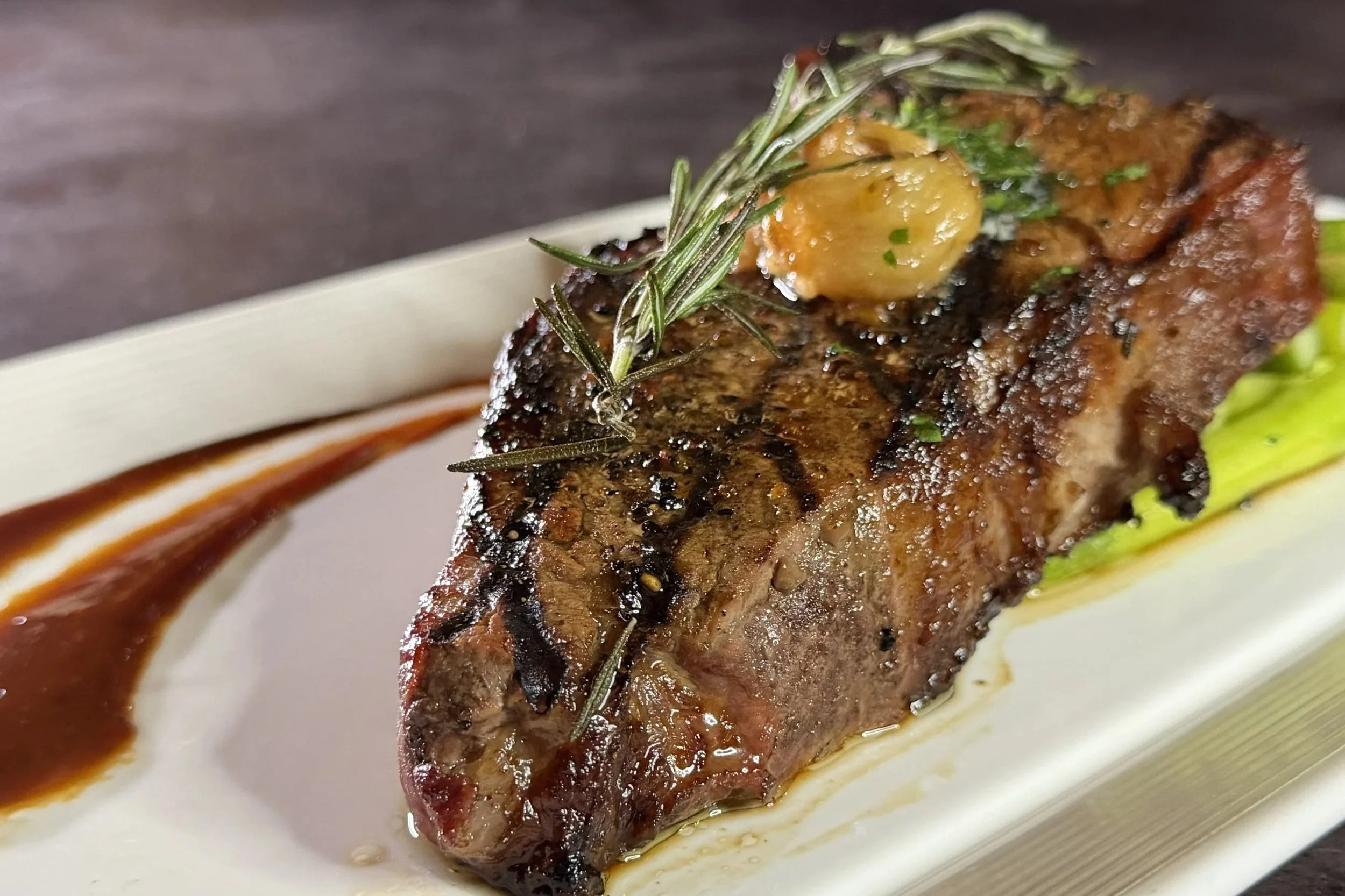 A grilled steak topped with a sprig of rosemary and a small dollop of garlic butter, served on a white rectangular plate with sauce on the side.