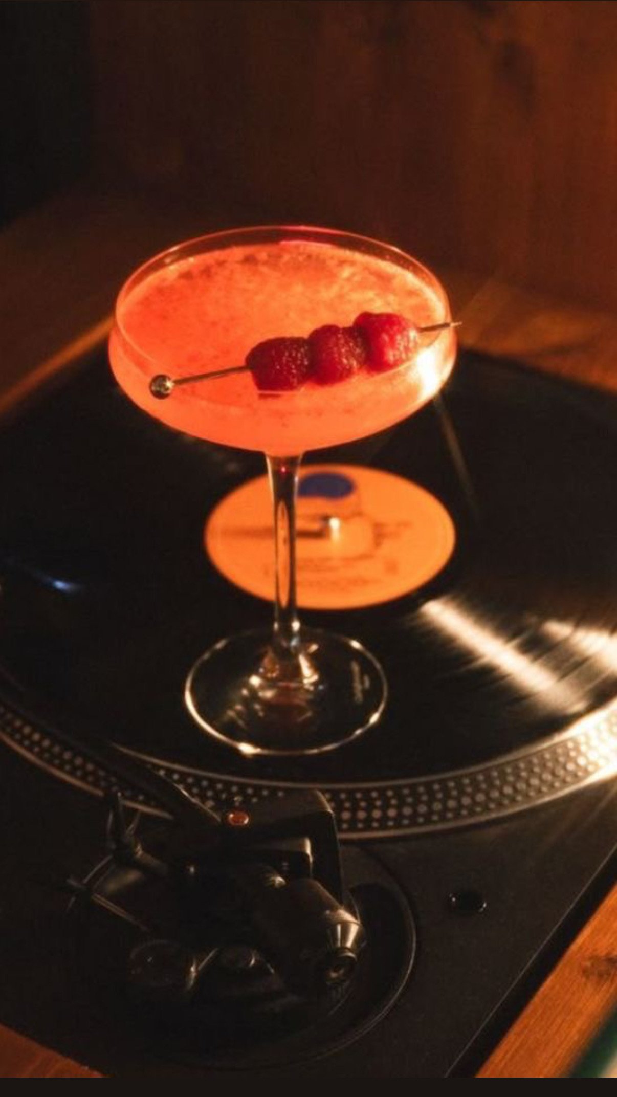 A pink cocktail with raspberries on a skewer sits on a turntable in a dimly lit environment.