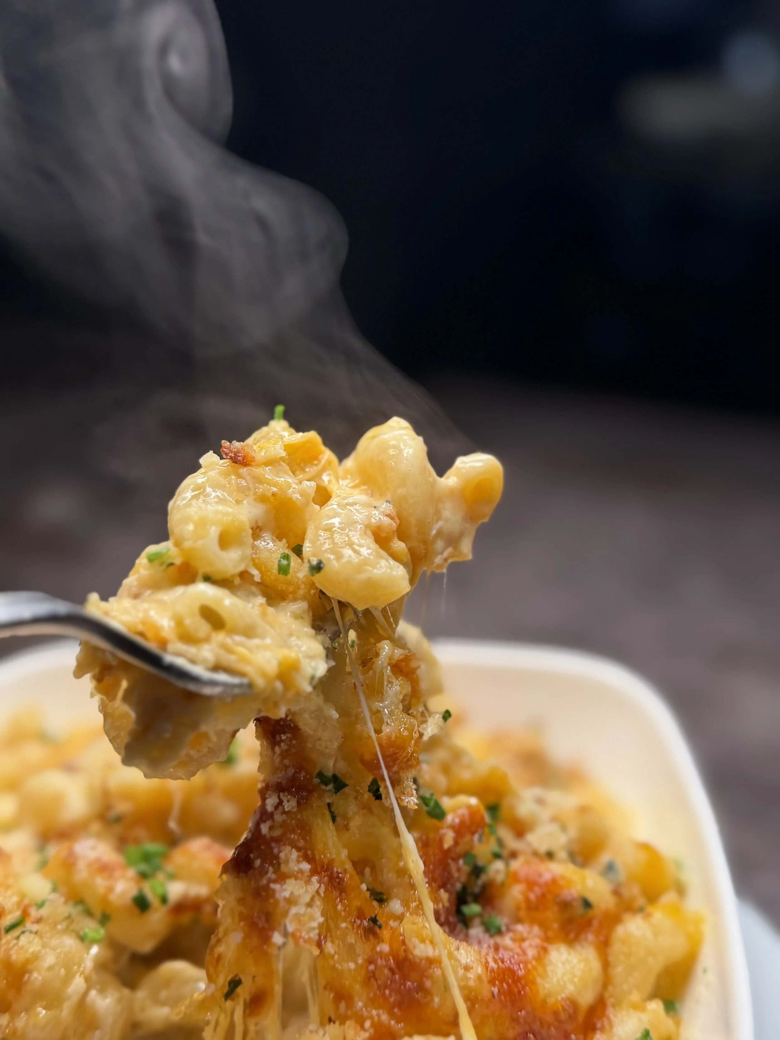 A fork lifting a bite of baked macaroni and cheese, with melted cheese strings, garnished with chopped chives, in a white dish.