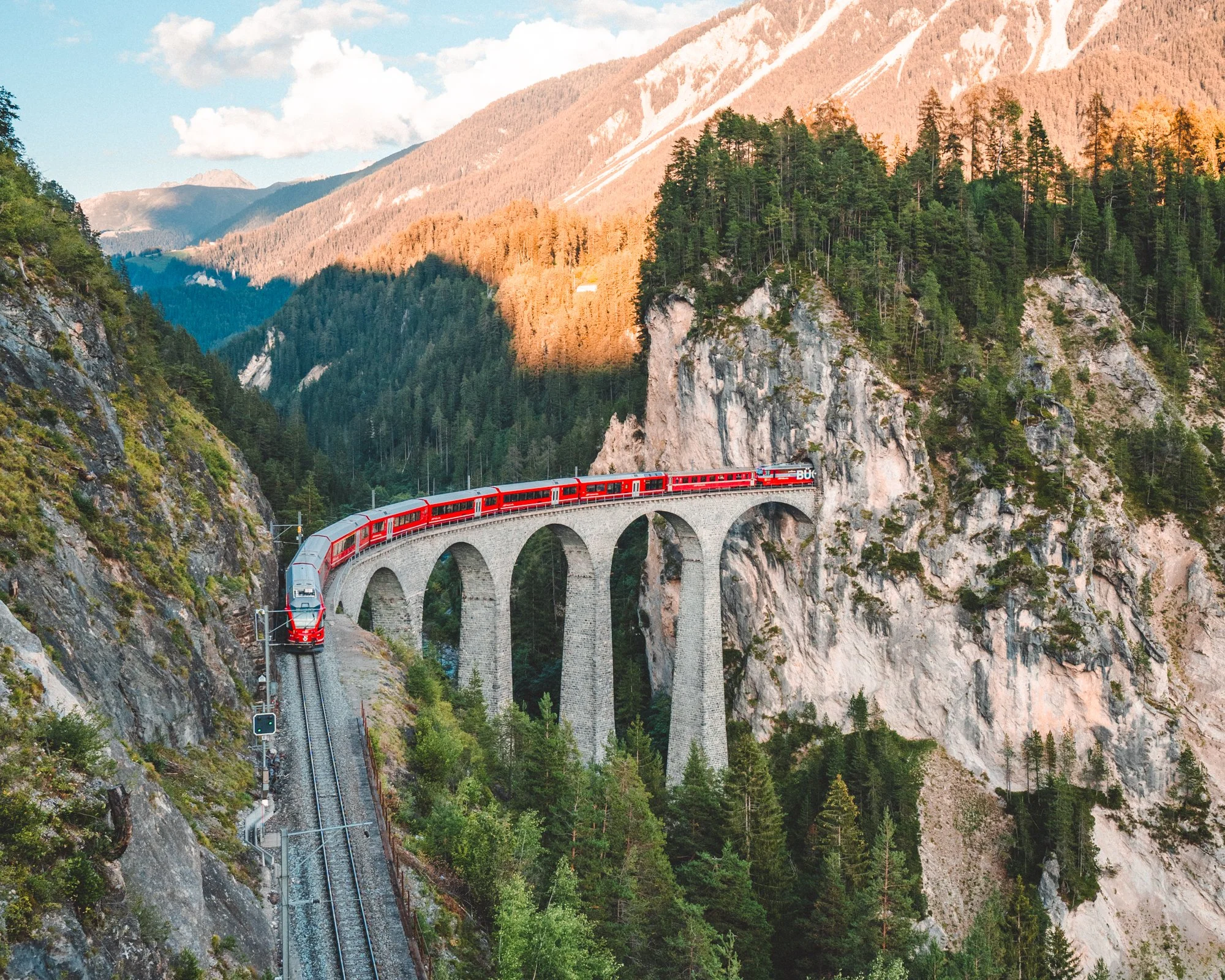 Trem de passageiros vermelho atravessando uma ponte de pedra em uma paisagem montanhosa com florestas de árvores verdes e rochas.