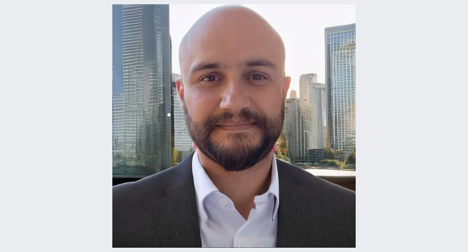 Homem de cabelo raspado, barba e bigode, usando terno escuro e camisa branca, posando na frente de uma janela com vista para edifícios altos de uma cidade moderna ao fundo.