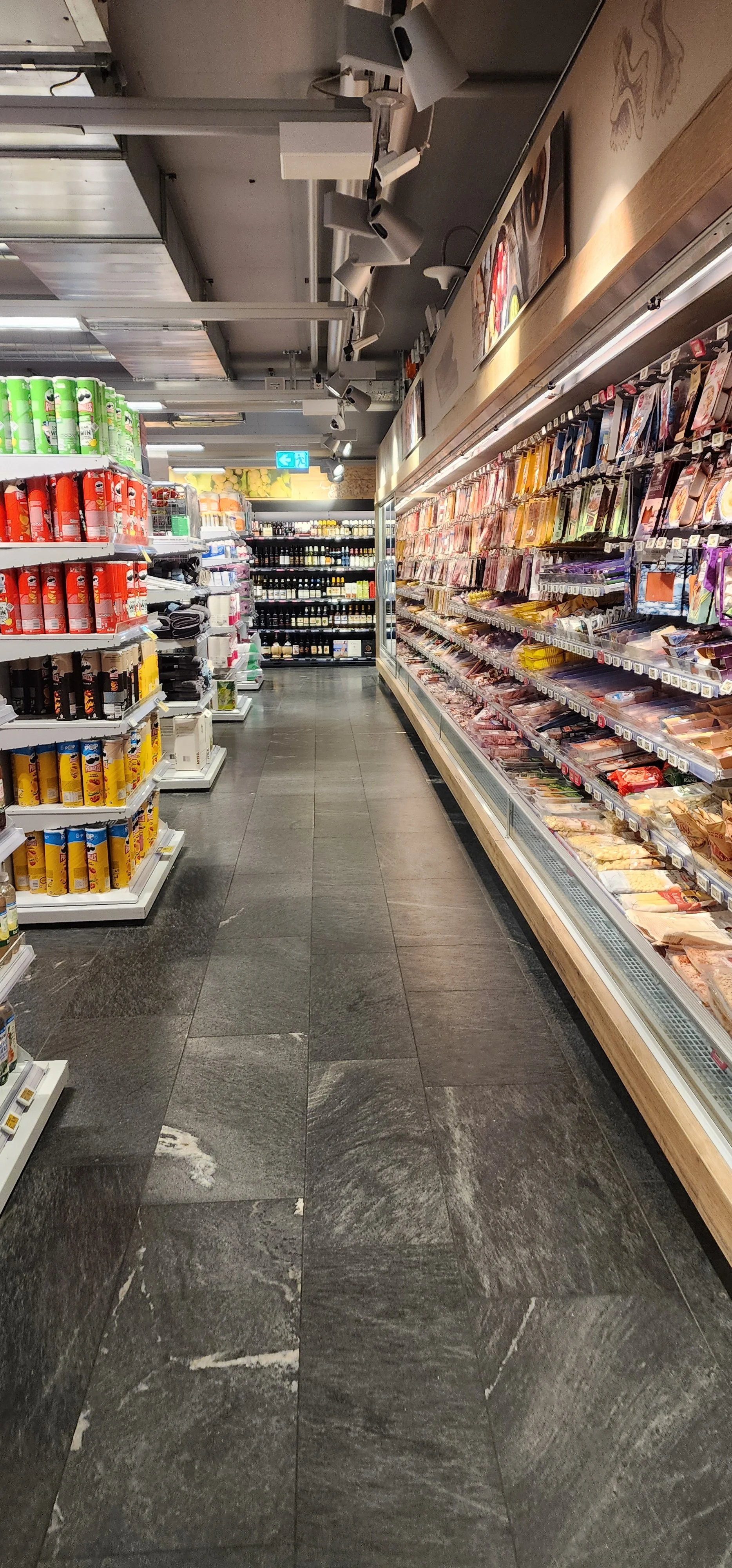 Corredor de supermercado com prateleiras cheias de produtos alimentícios e itens de consumo, piso de azulejos escuros, iluminação de teto com foco nos produtos, saída de emergência visível ao fundo.