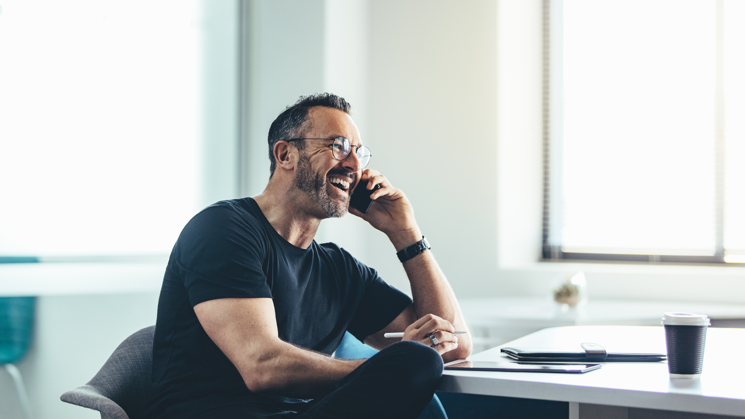 A man with glasses and a beard is smiling while talking on his phone in an office, sitting at a desk with a laptop, a tablet, a pen, and a coffee cup.