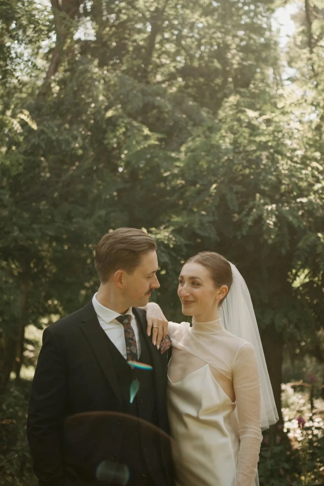 Ein heiratendes Paar in einem Park, die Frau trägt ein weißes Kleid und einen Schleier, der Mann trägt einen dunklen Anzug.