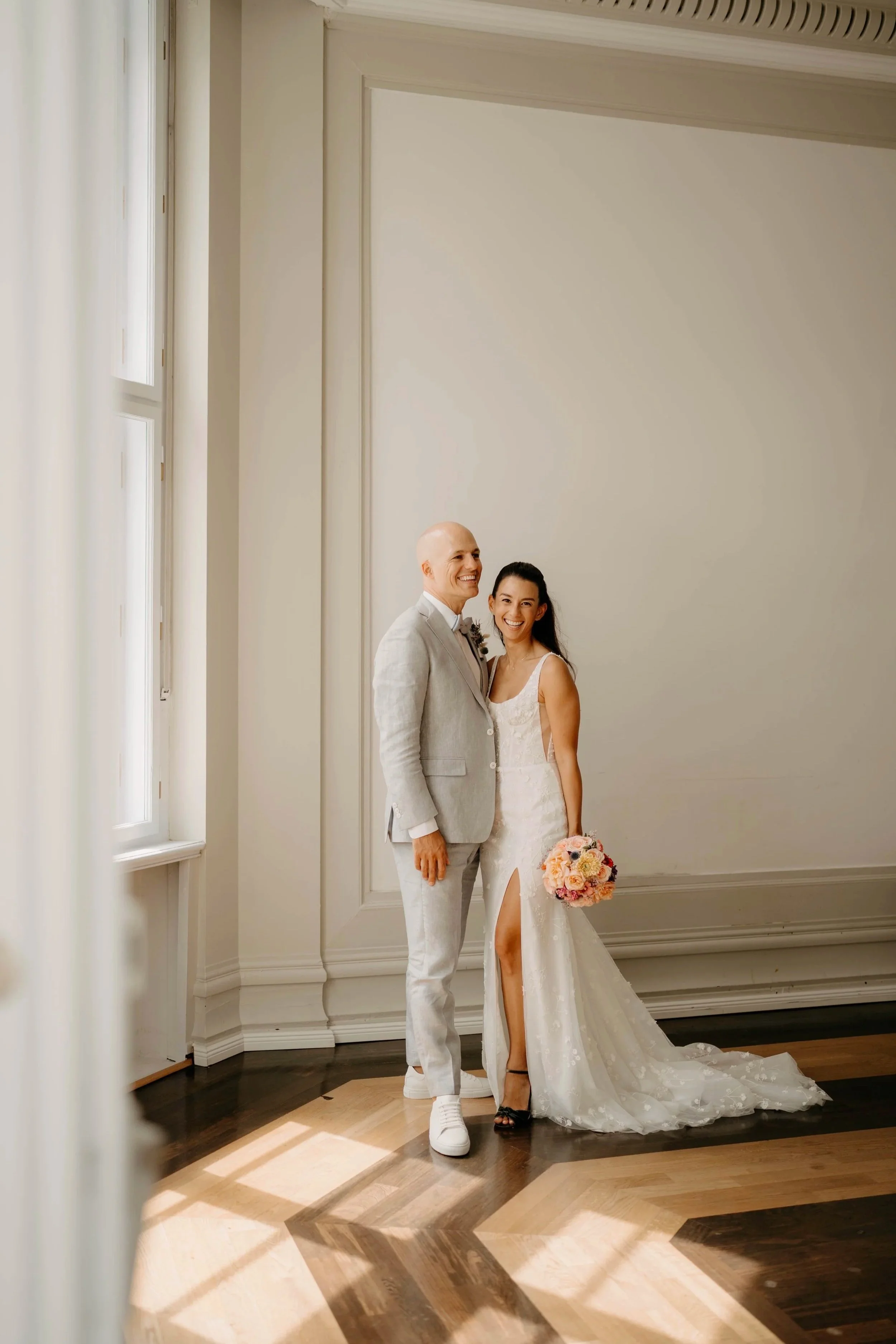 Ein Brautpaar in Hochzeitskleidung posiert vor einer leeren weißen Wand, die natürliche Beleuchtung durch Fenster auf der linken Seite.
