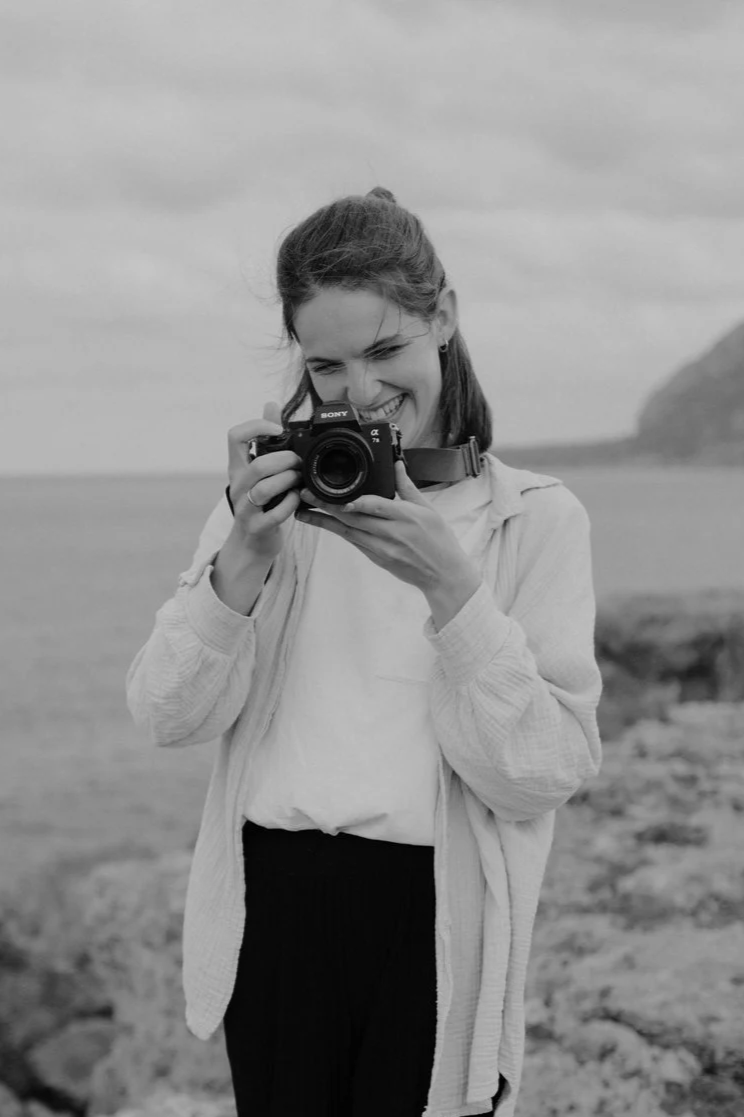 Eine lachende junge Frau in einem weißen Pullover und dunklen Hosen, die eine Kamera in der Hand hält, fotografiert im Freien an einem Strand oder einer Küstenlandschaft.