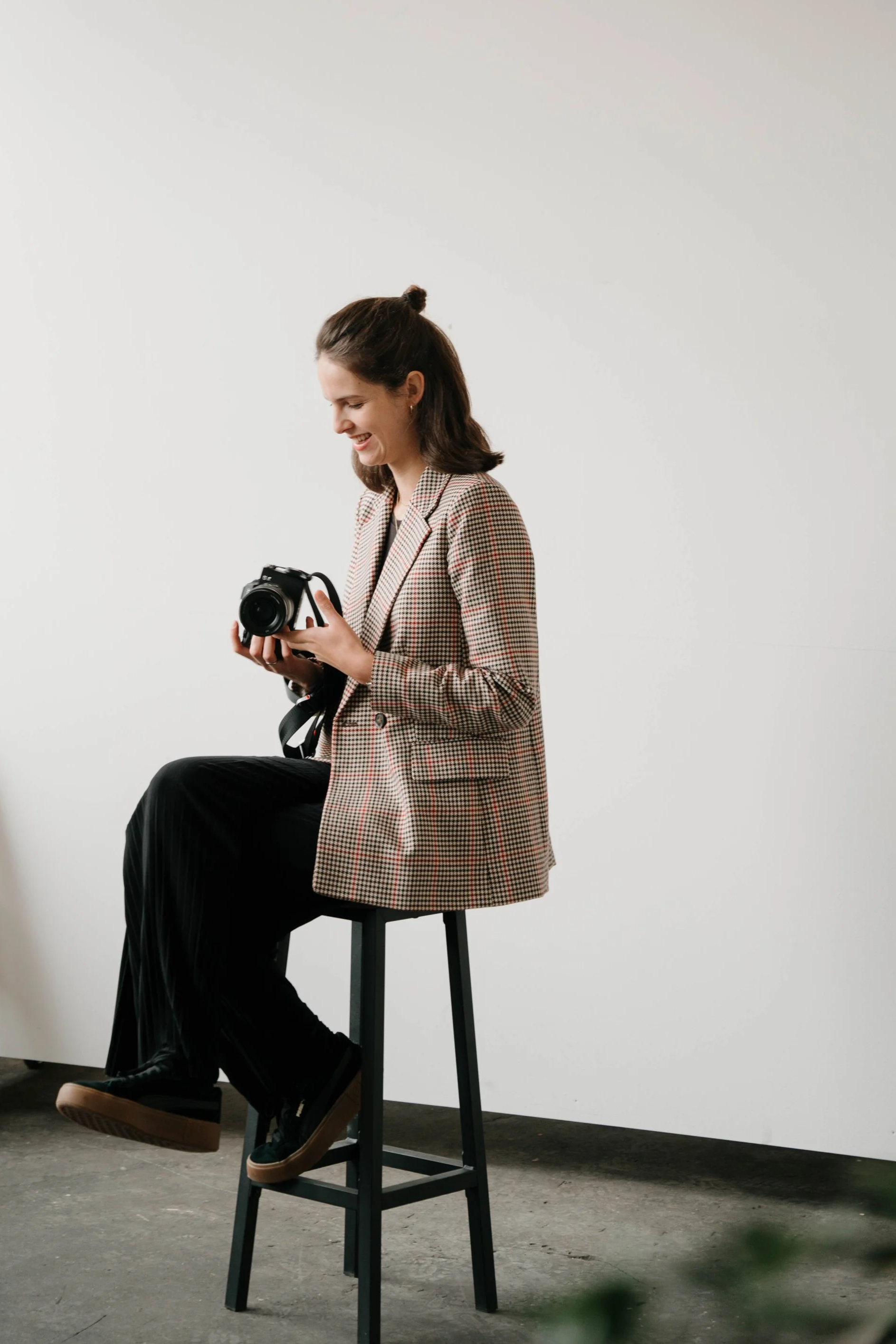 Eine Frau sitzt auf einem hohen Stuhl vor einer weißen Wand und hält eine Kamera in den Händen, lächelnd.