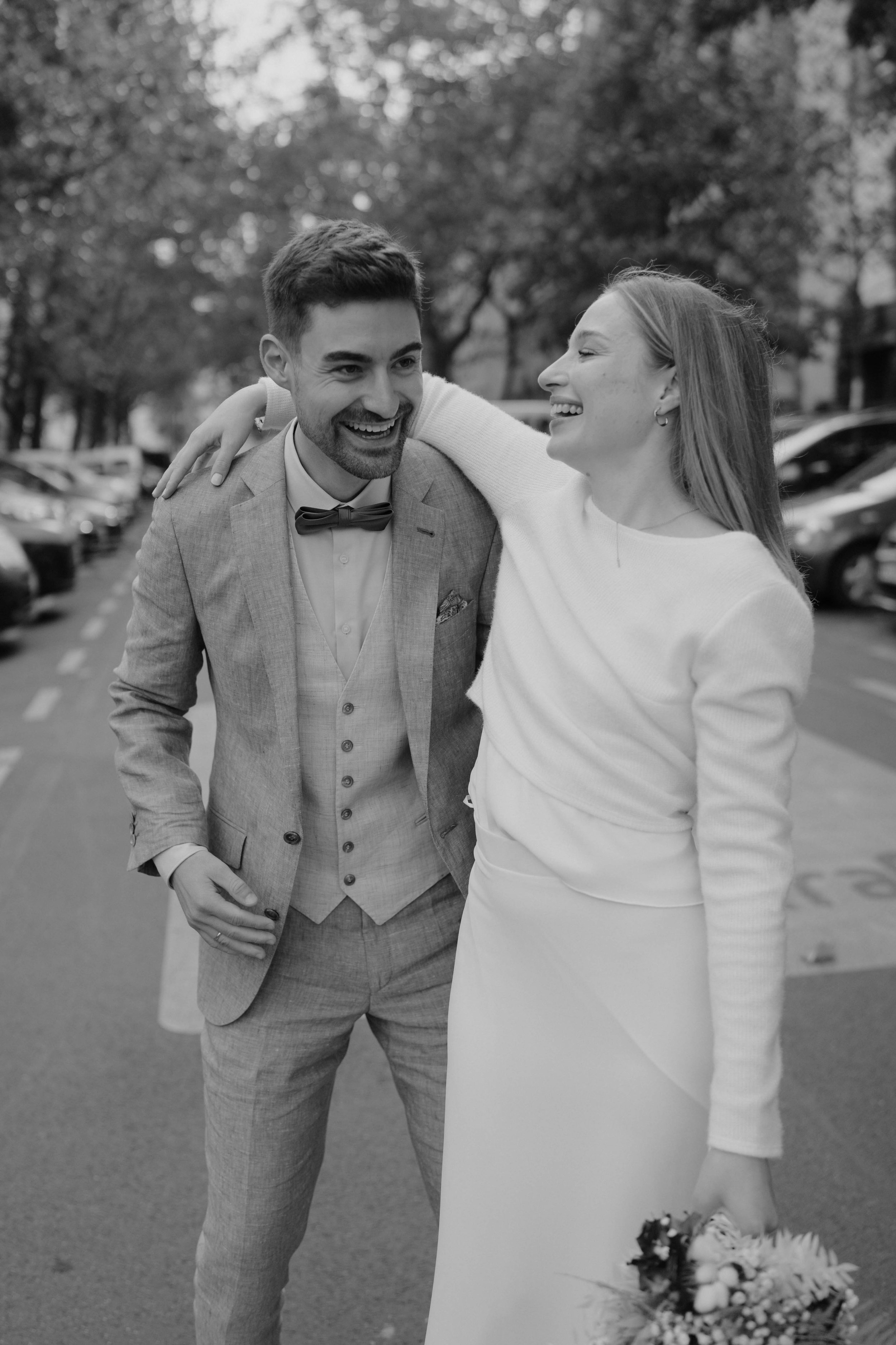 Zwei Menschen, ein Mann in Anzug und eine Frau in einem weißen Kleid, lachen und umarmen sich auf einer Straße, vermutlich bei einer Hochzeit.