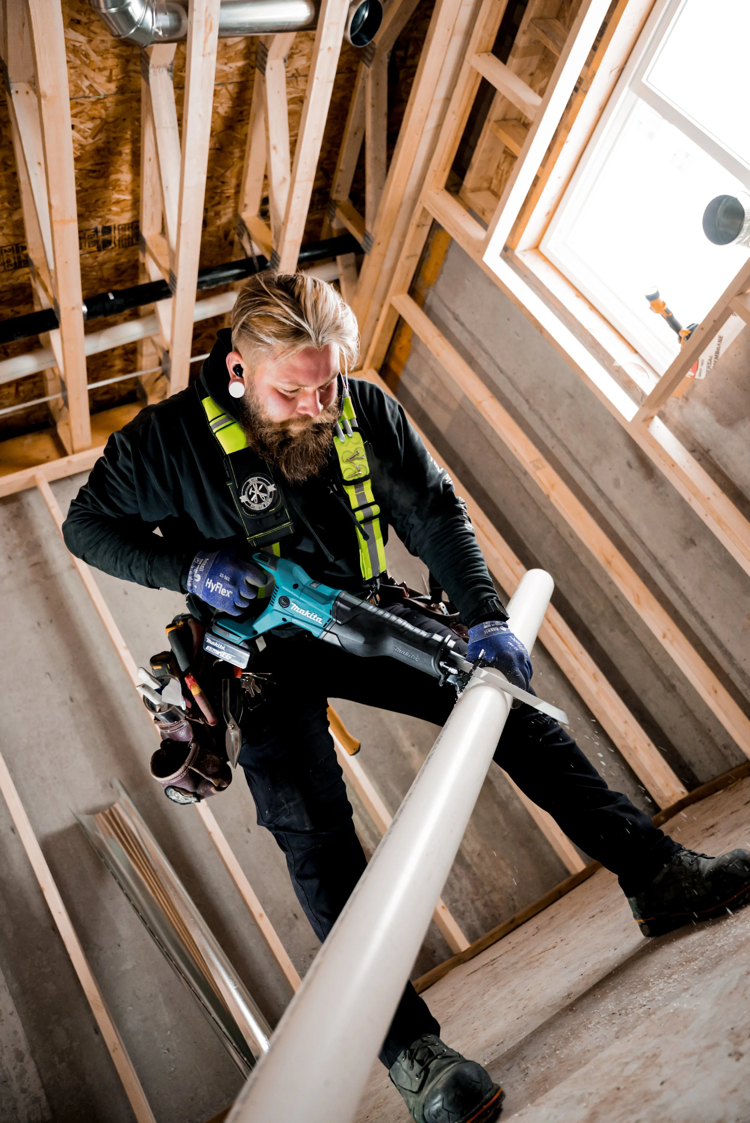 A construction worker with a beard and earrings is cutting a pipe with a power saw inside a building under construction, with exposed wooden framing and a window.