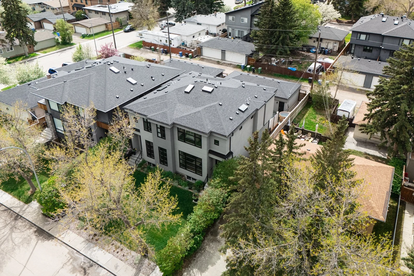 Aerial view of a modern multi-unit residential building with a gray roof, surrounded by a small green yard with trees, in a neighborhood with similar houses.