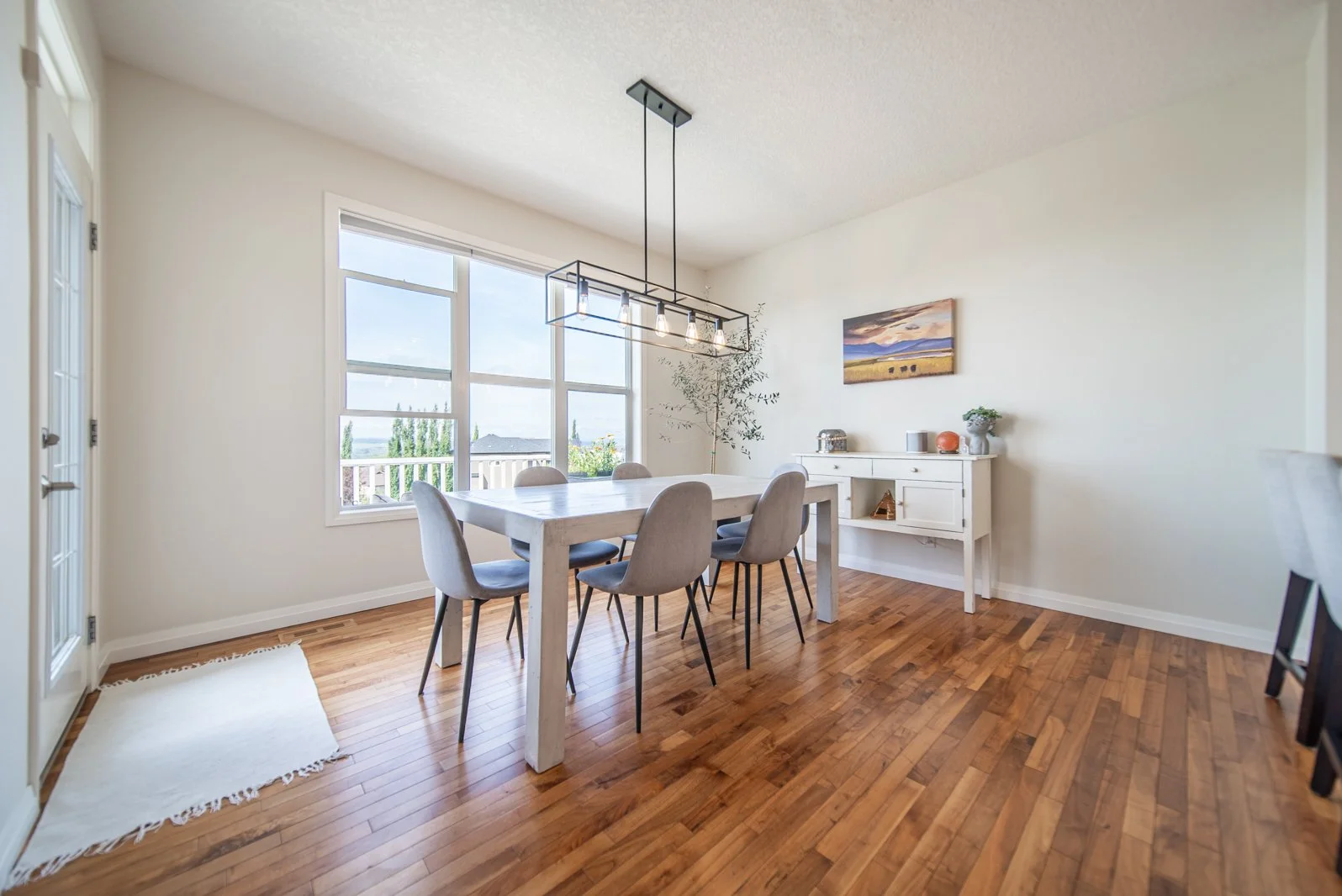 Dining room with large window, white table, six gray chairs, hardwood floors, white sideboard with decorative items, painting, and a pendant light fixture.