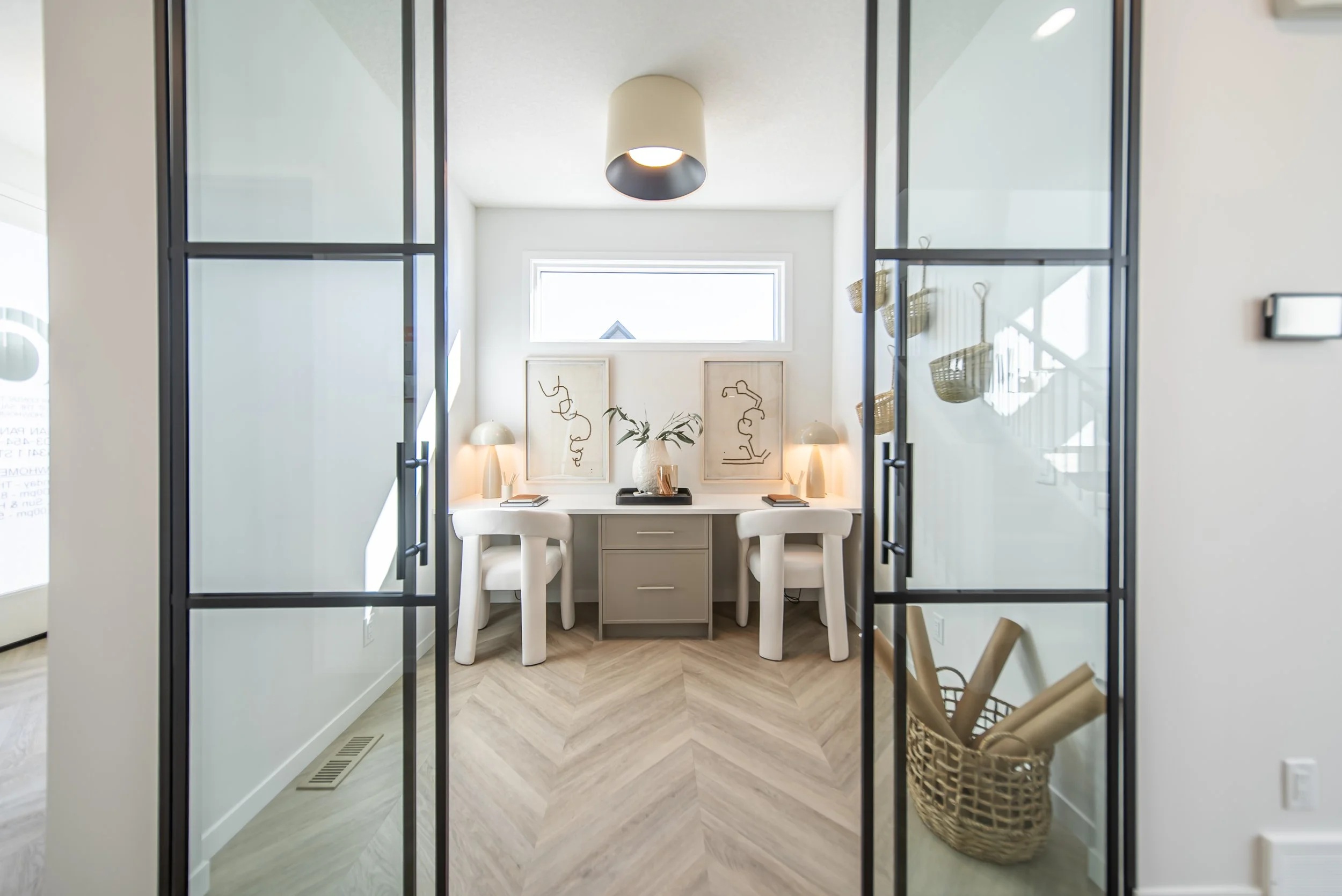 Home office space with two white chairs at a white desk, framed abstract art, and wicker baskets on the right, enclosed by glass doors.